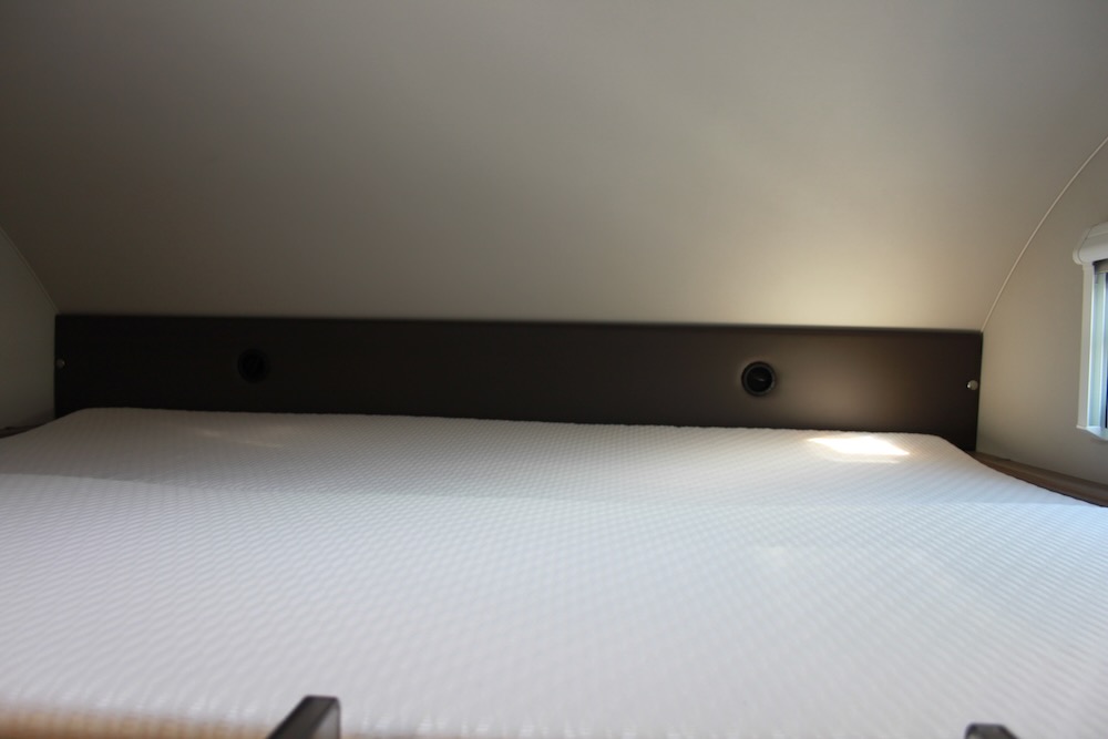 Cozy over-cab sleeping area in an RV rental, featuring a white mattress, dark panel, and a window with blinds.. Knaus Live Wave 600 DKG 2021