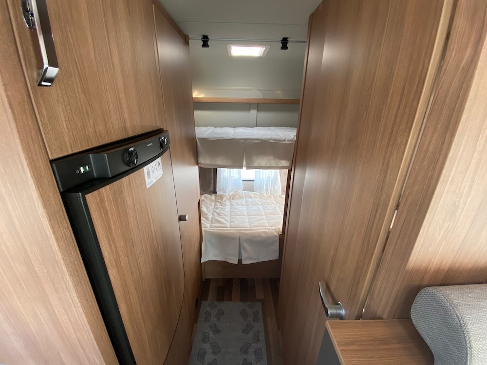 Compact RV rental interior featuring a refrigerator on the left and two bunk beds down a narrow aisle, showcasing efficient design.. Weinsberg Cara Home 600 DKG 2022