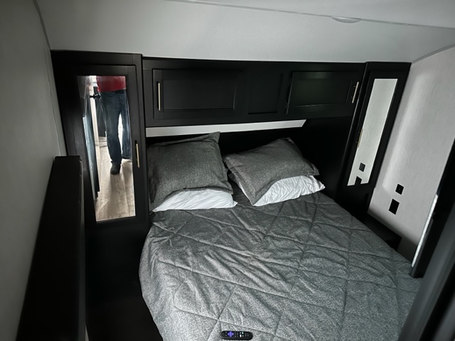 Compact RV rental bedroom with a gray bed, dark overhead storage, and a mirrored closet door on the left.. Forest River Cherokee 2022