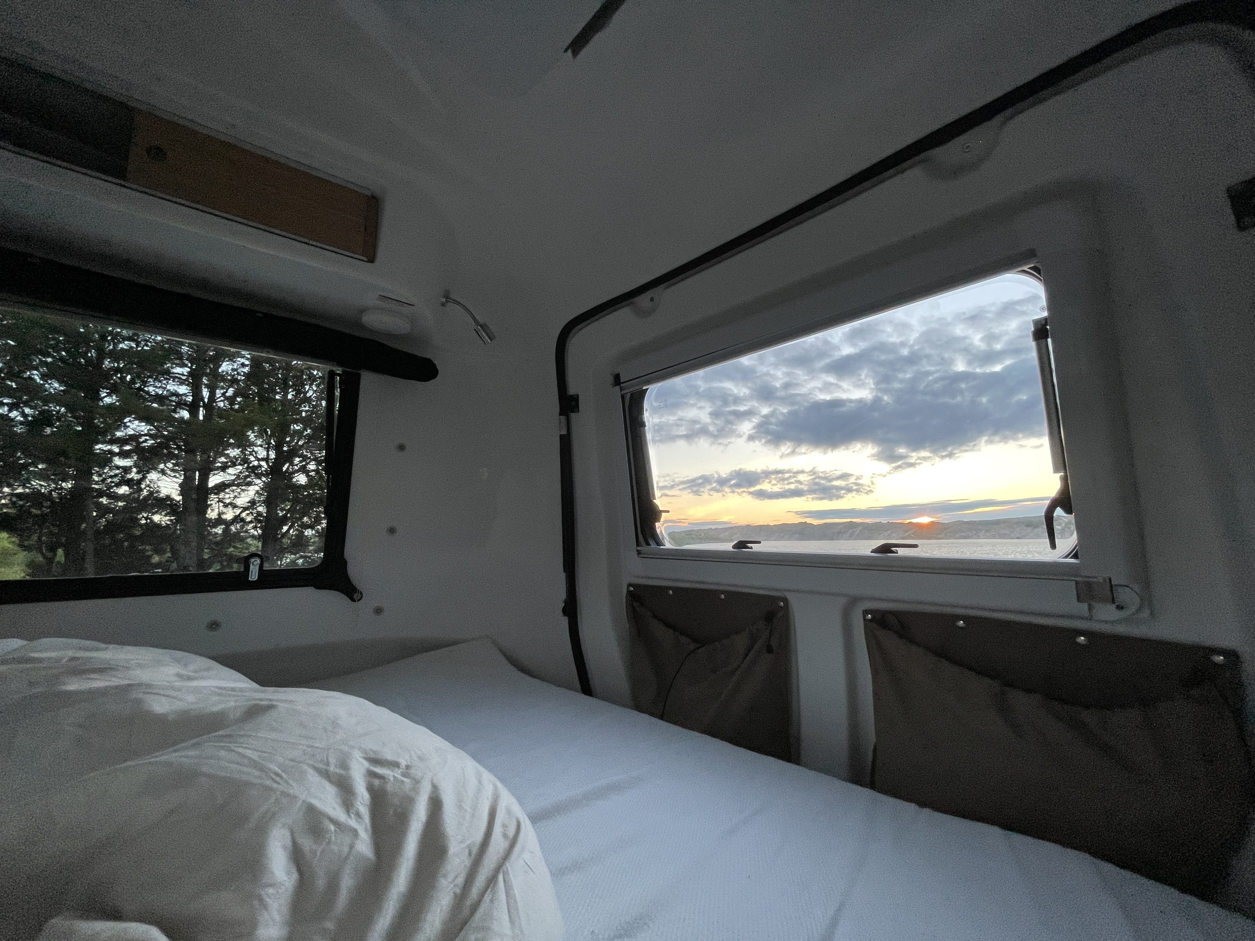 View from inside an RV rental (camper): a cozy bed and two windows, one showing a sunset over water, the other trees.. Happier Camper HC1 2021