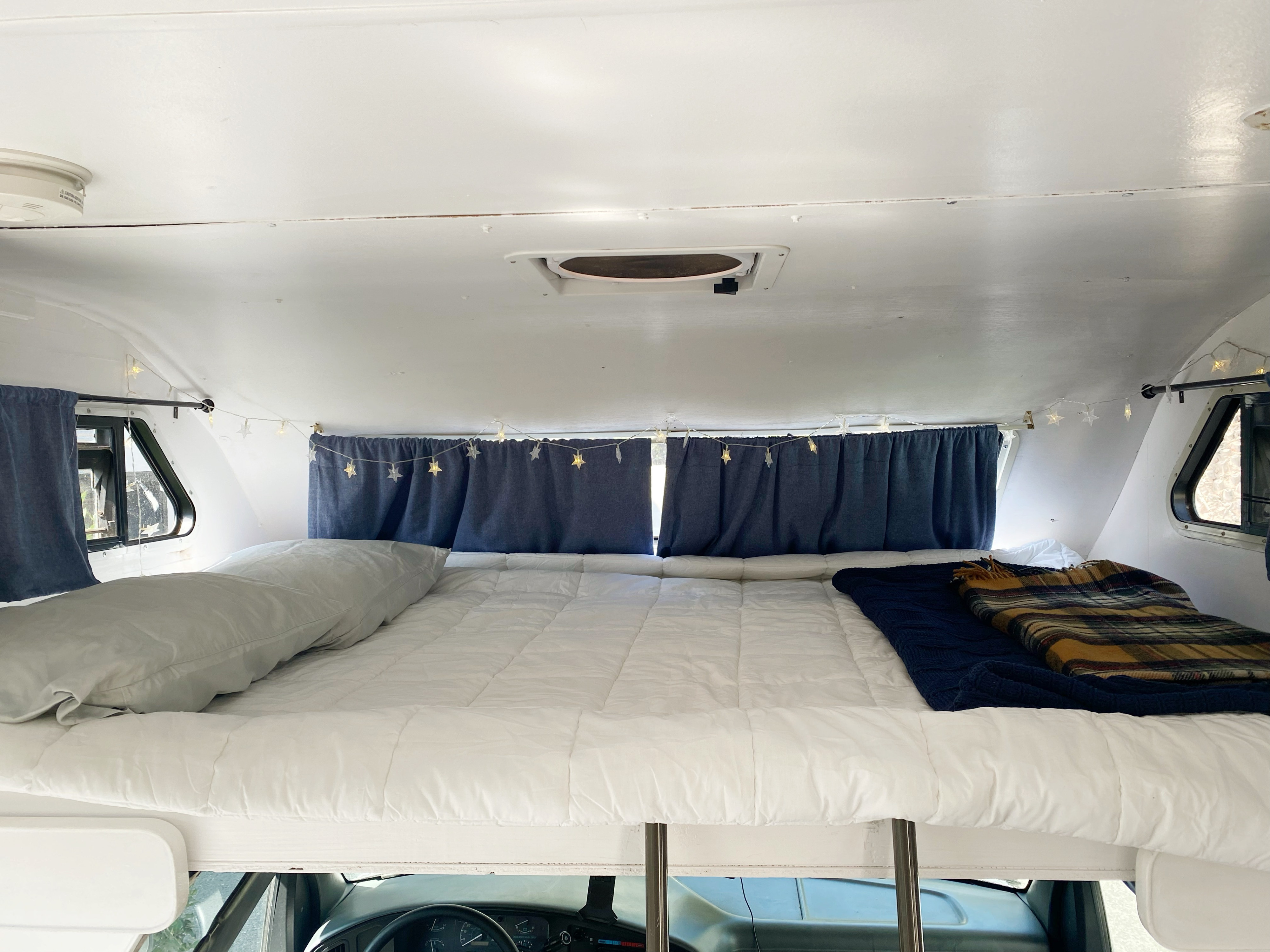 Cozy RV rental cab-over bed with white bedding, blue curtains, and star lights for a comfortable road trip sleep setup.. Ford Jamboree 1992