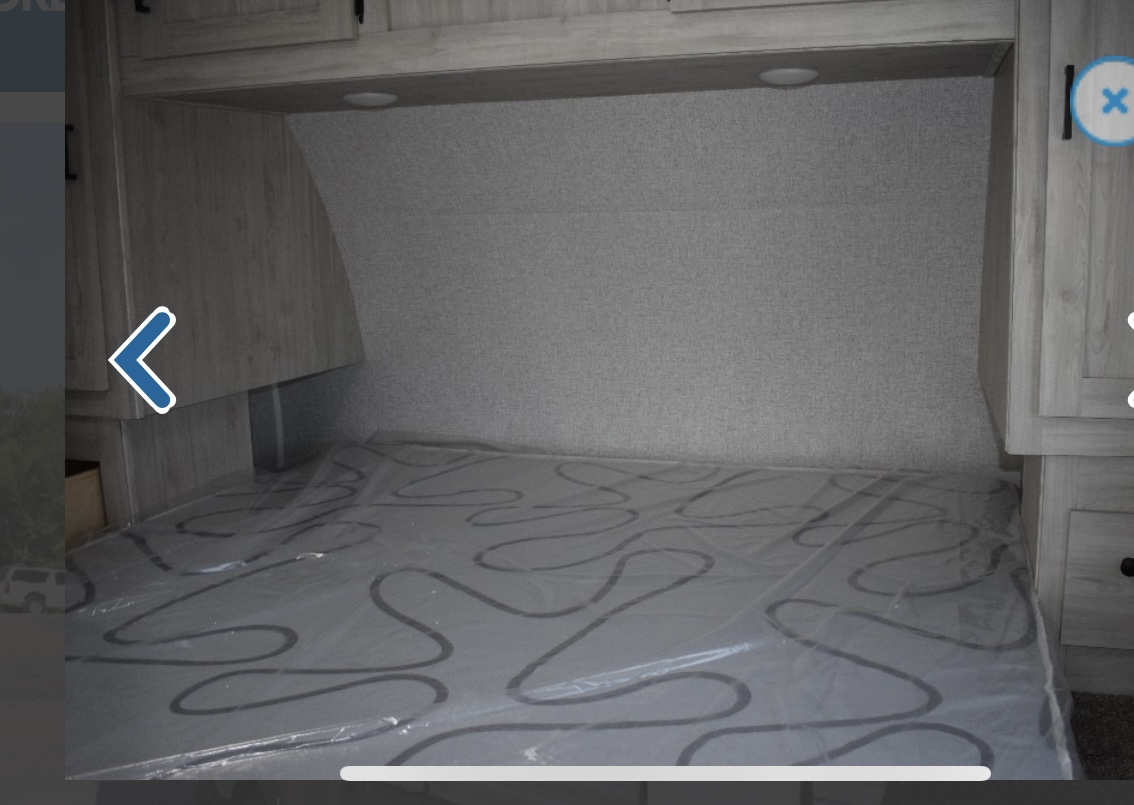 Interior of an RV rental bedroom showing a new, plastic-wrapped mattress, overhead storage, and bedside cabinets.. East to West Silver Lake 2022