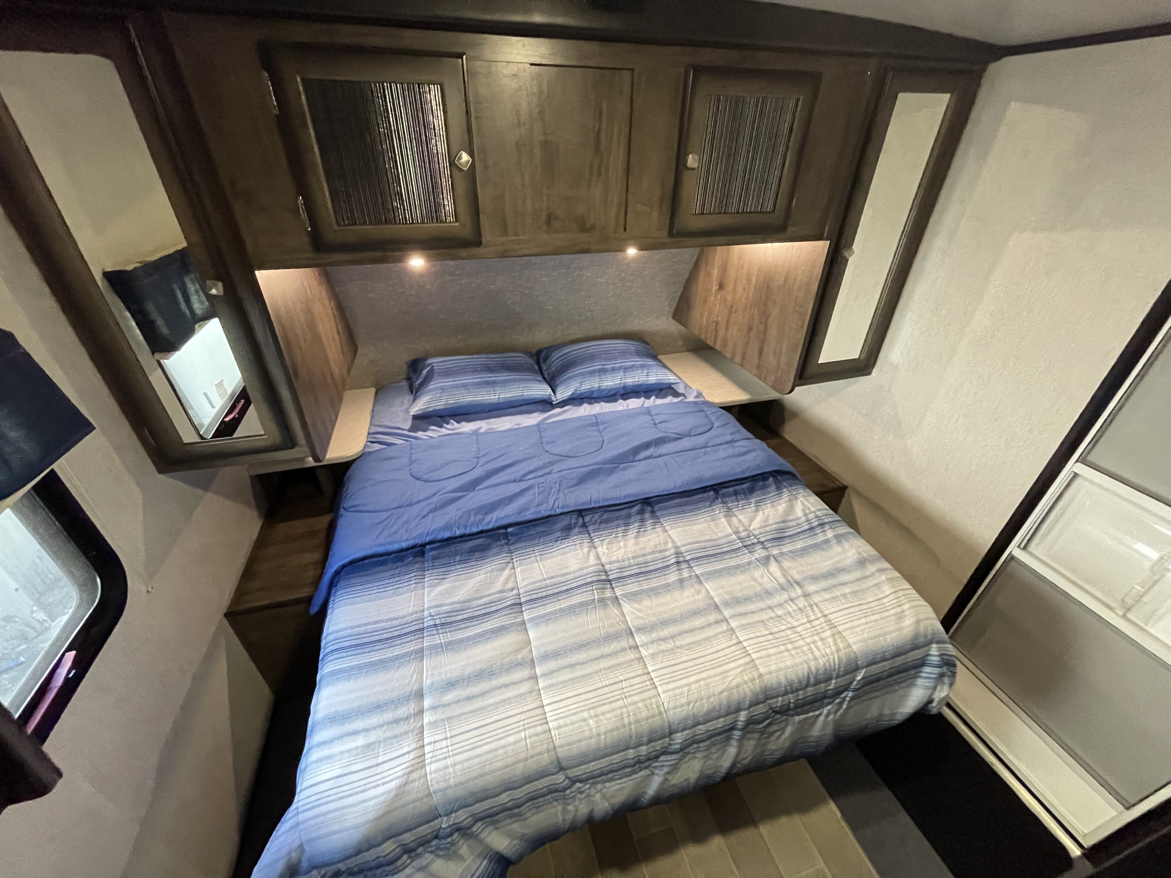 Cozy RV rental bedroom featuring a striped bed, overhead cabinets with lights, and side shelves.. Heartland RVs Mallard 2019