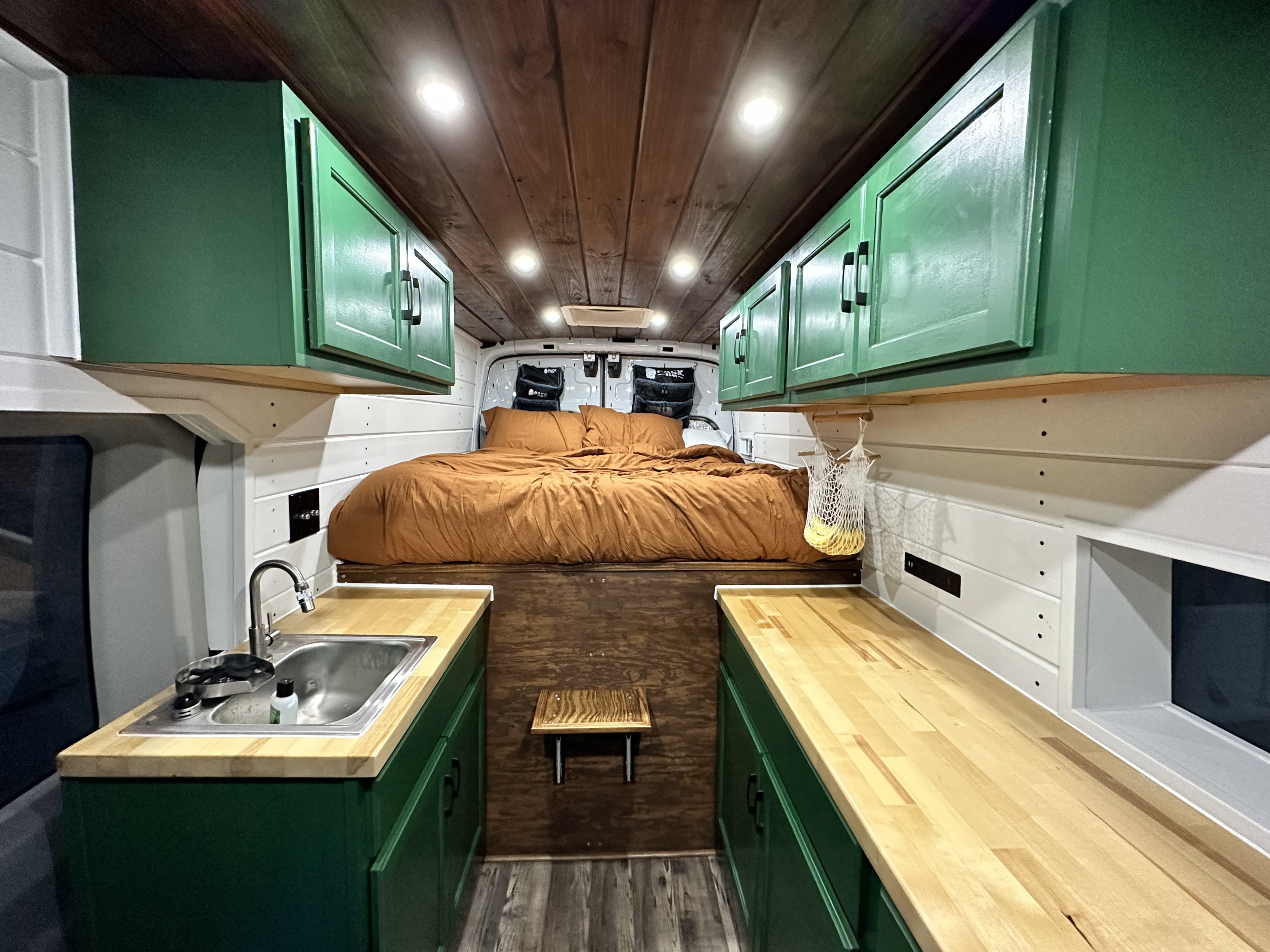 Cozy RV rental interior with a bed, wood accents, kitchen sink, and green overhead cabinets.. Ford Transit 2015