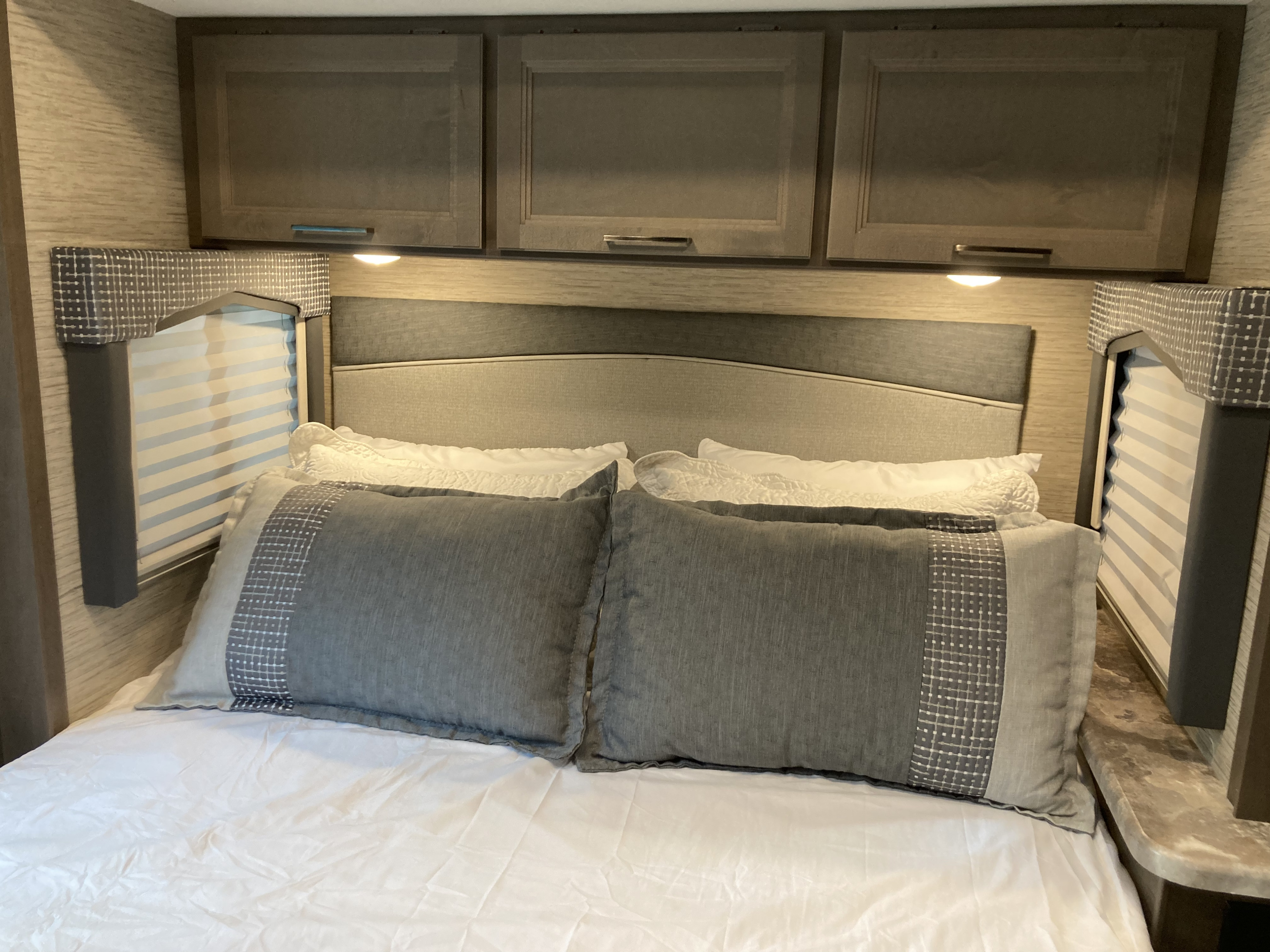 Cozy RV rental bedroom with upholstered headboard, crisp white sheets, grey decorative pillows, and modern overhead storage.. Thor Motor Coach Chateau 2023