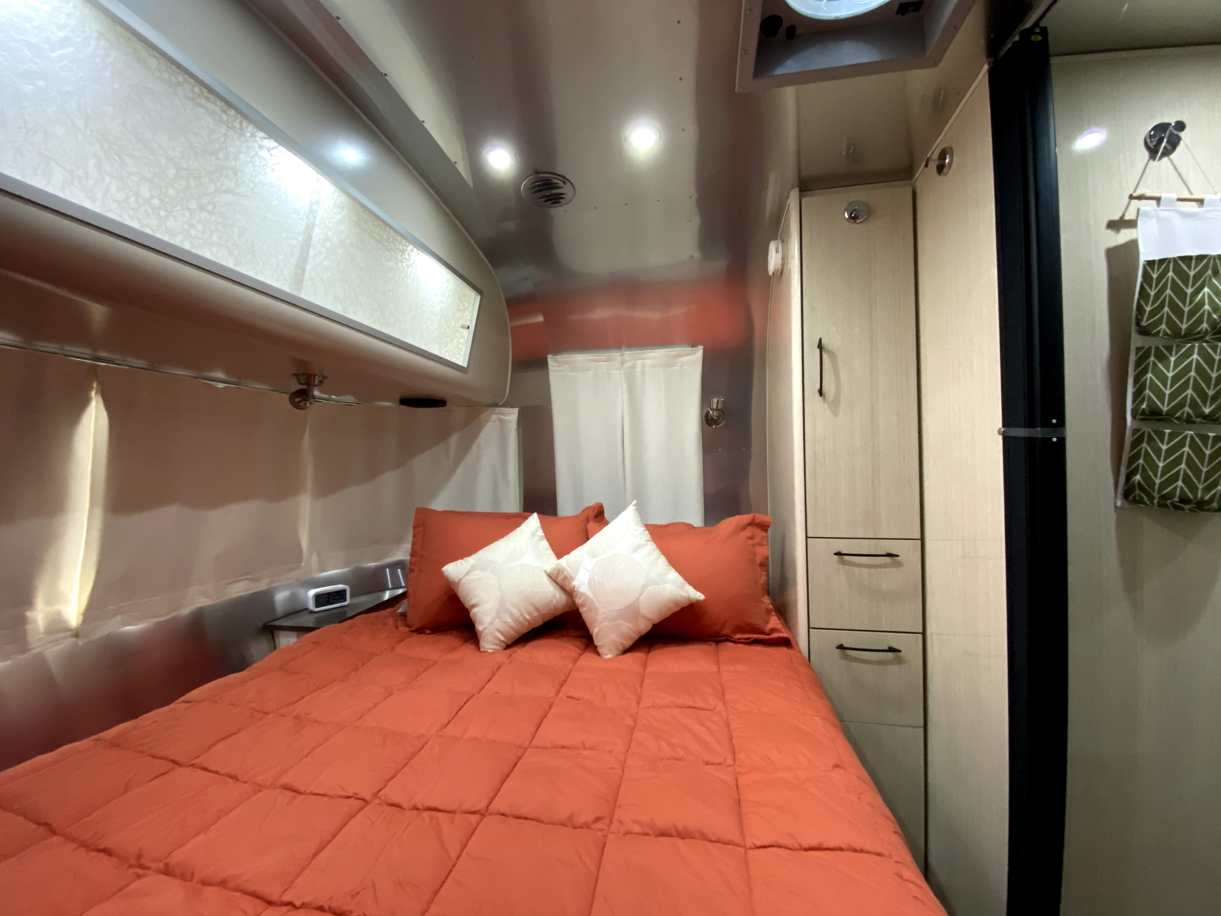 Step inside this cozy RV rental's modern bedroom. Features include an orange bed, shiny aluminum walls, and compact storage.. Airstream International 2017
