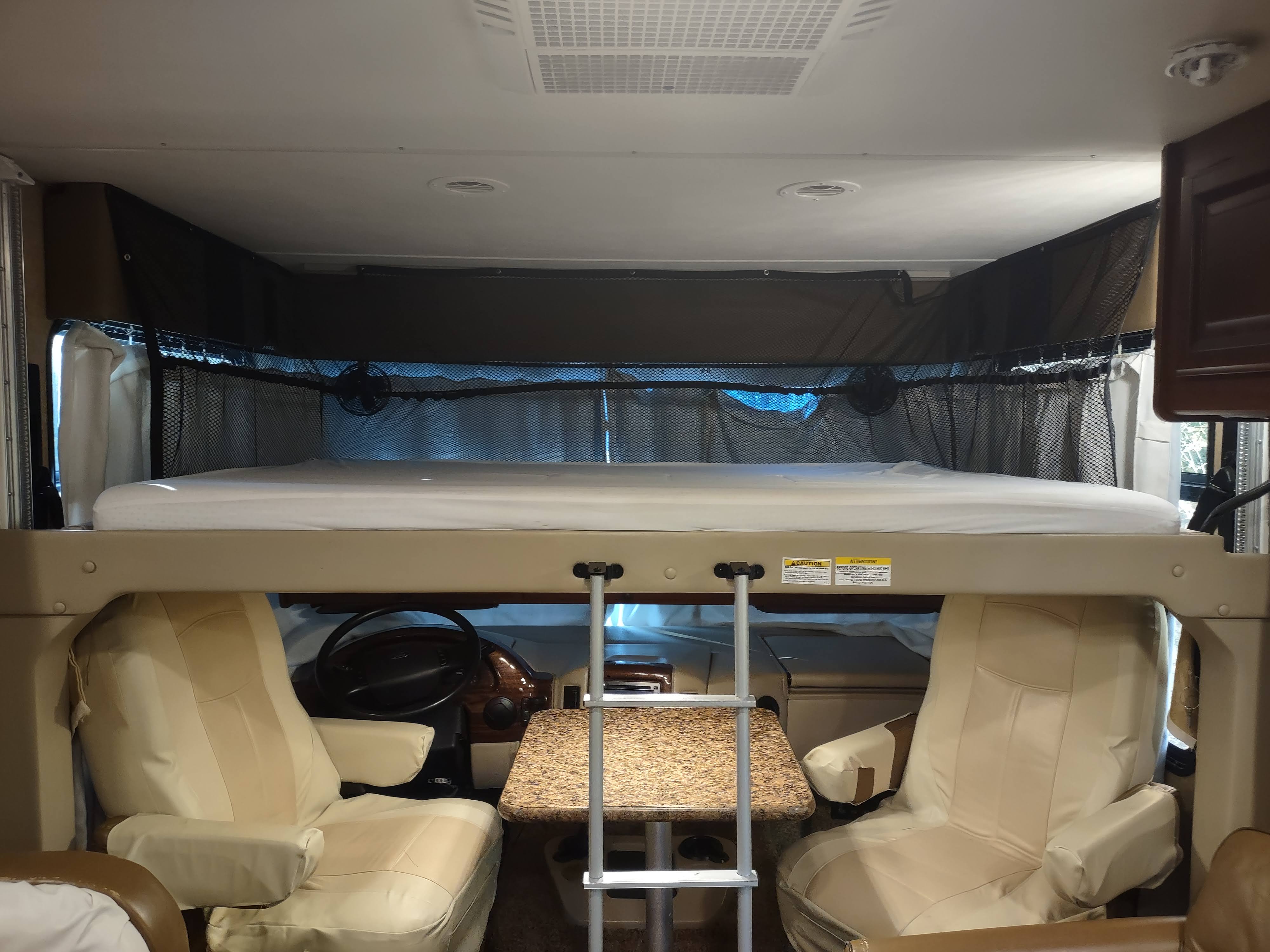An RV rental's over-cab bunk bed with safety netting, positioned above the driver and passenger seats, ready for travel.. Thor Windsport 2015