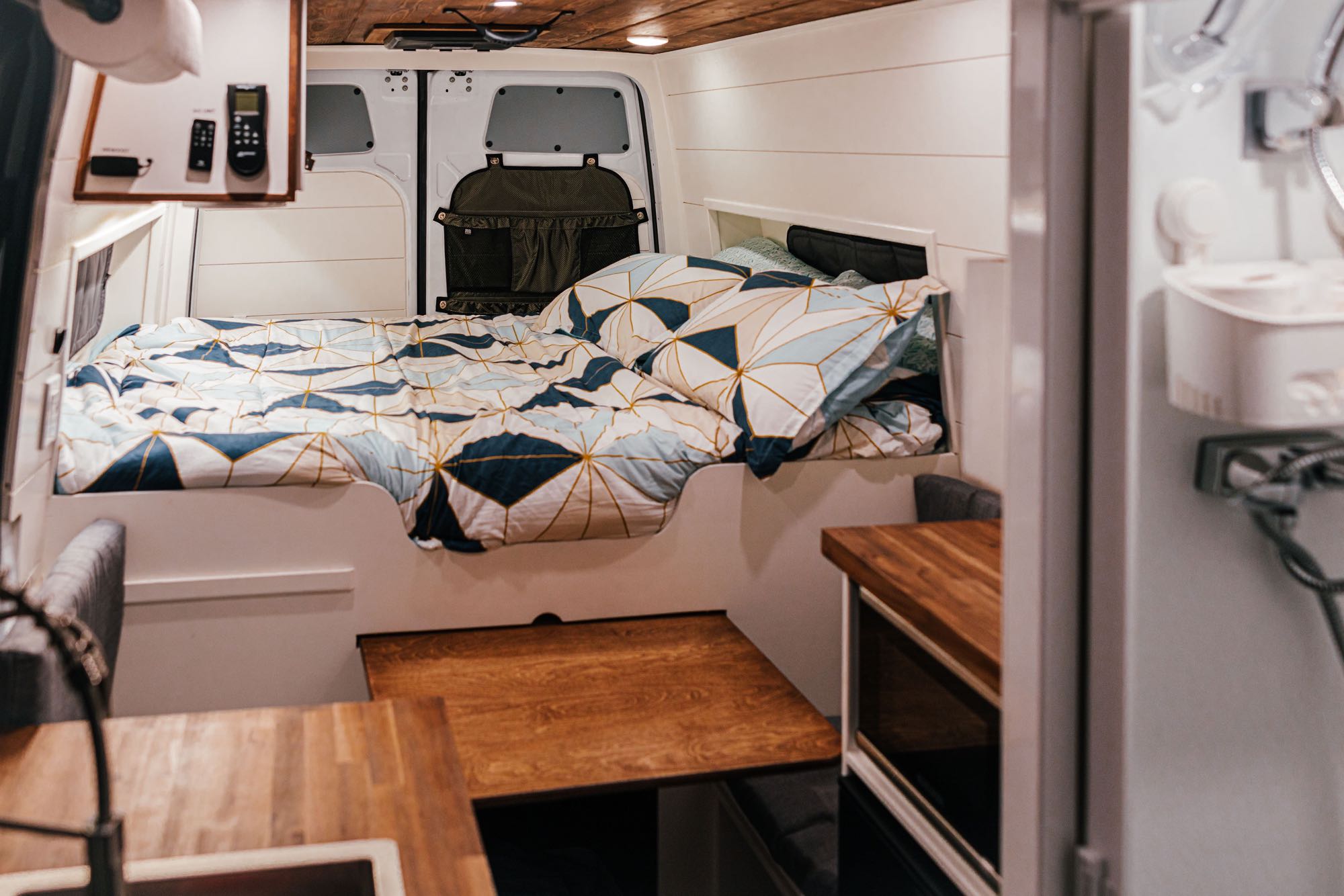 Inside a modern **rv rental**: a cozy bed with patterned duvet, white walls, and wooden table, showing efficient van living.. Mercedes-Benz Sprinter RV Motorhome Campervan 2022