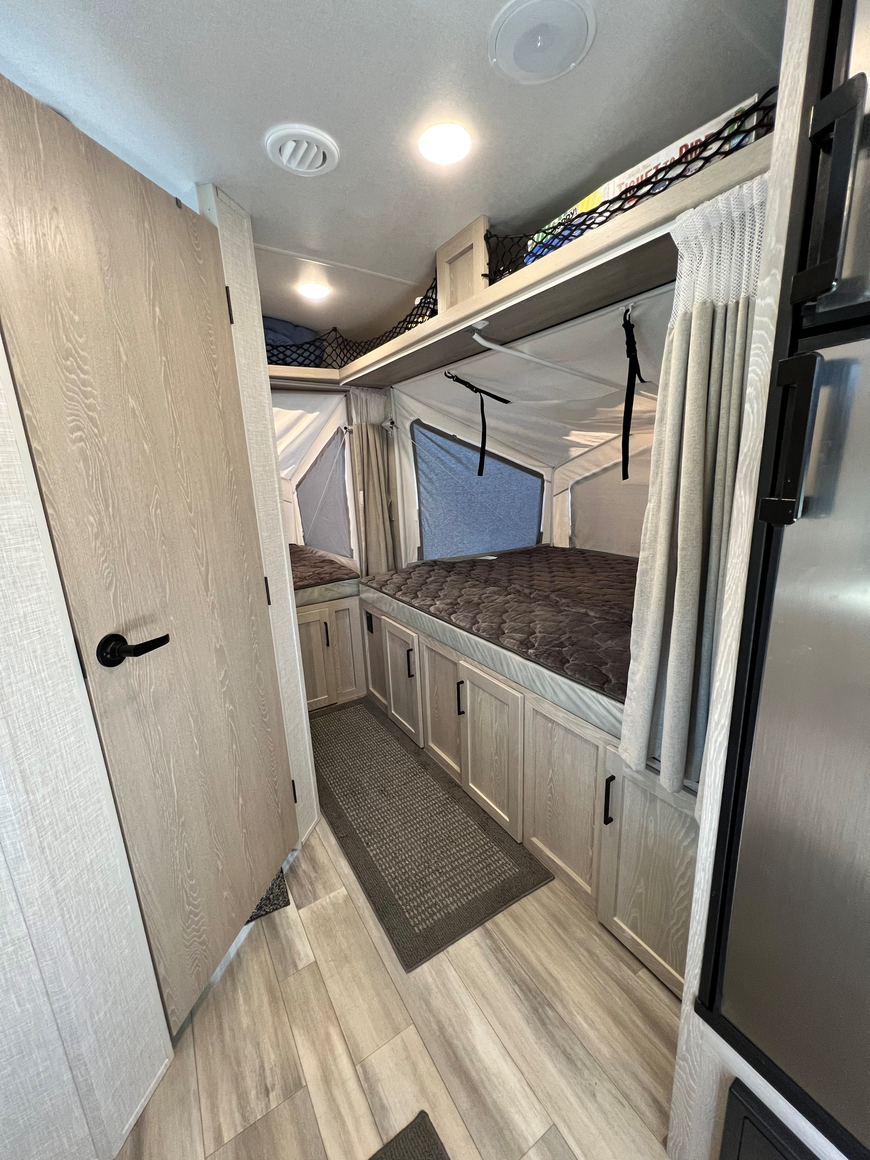 Bright RV rental interior with two beds, light wood cabinetry, overhead storage nets, and a hallway leading to another bed.. Forest River Rockwood Roo 2022