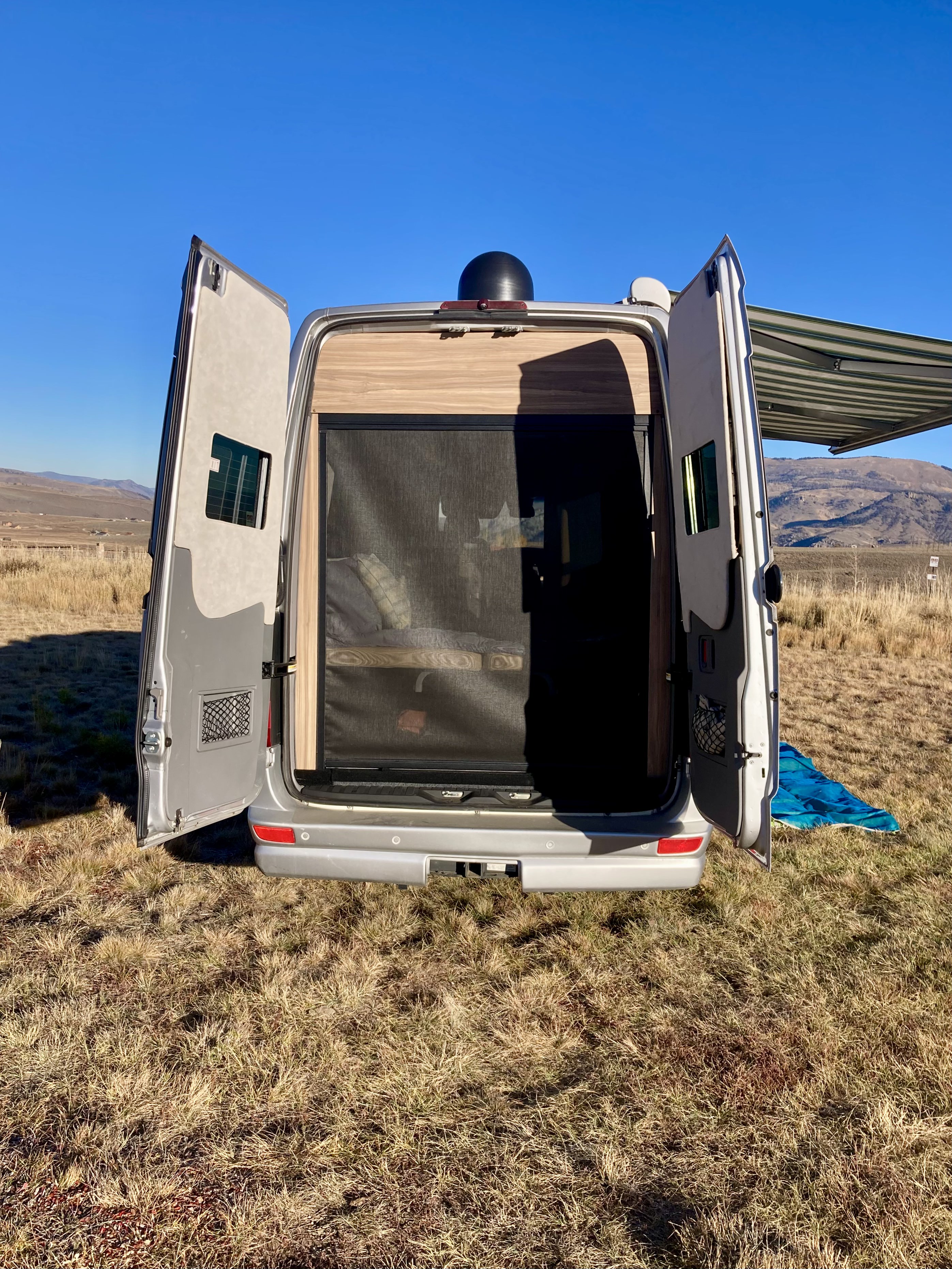 RV rental: Rear view of a silver camper van with open doors, screen, and awning, parked in a sunny, dry field.. Airstream Interstate 2017