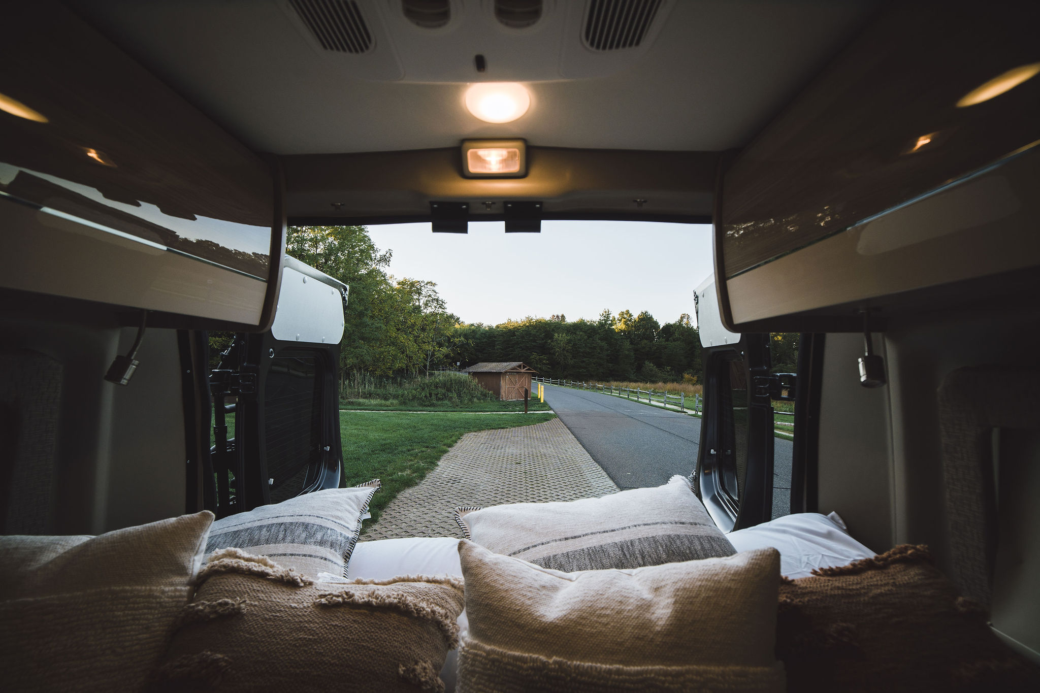 Inside a cozy RV rental: comfy bed, open doors revealing a scenic road, trees, and shed. Ready for adventure!. Thor Sequence 2022