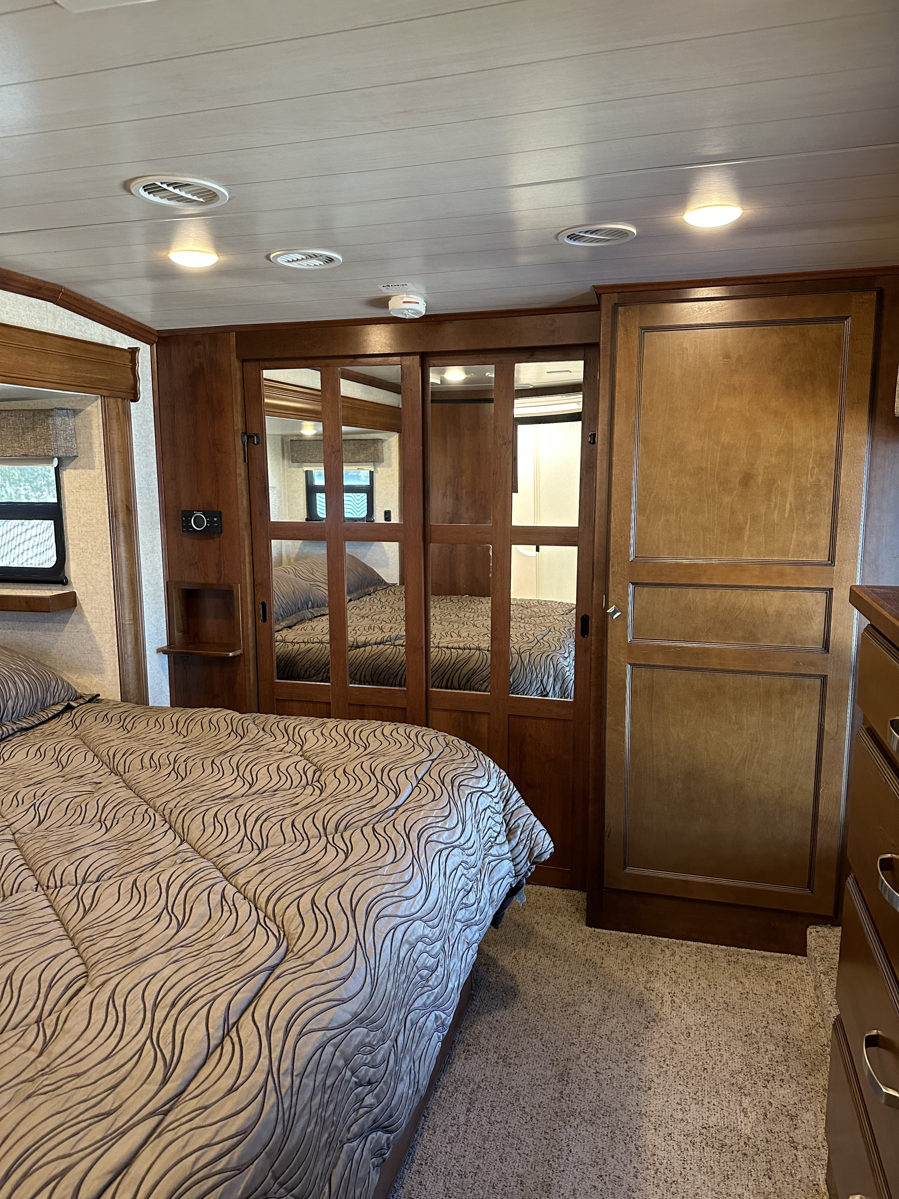 An RV rental bedroom featuring a patterned bed, mirrored wardrobe, wood cabinets, and ceiling vents.. Heartland Bighorn 2017
