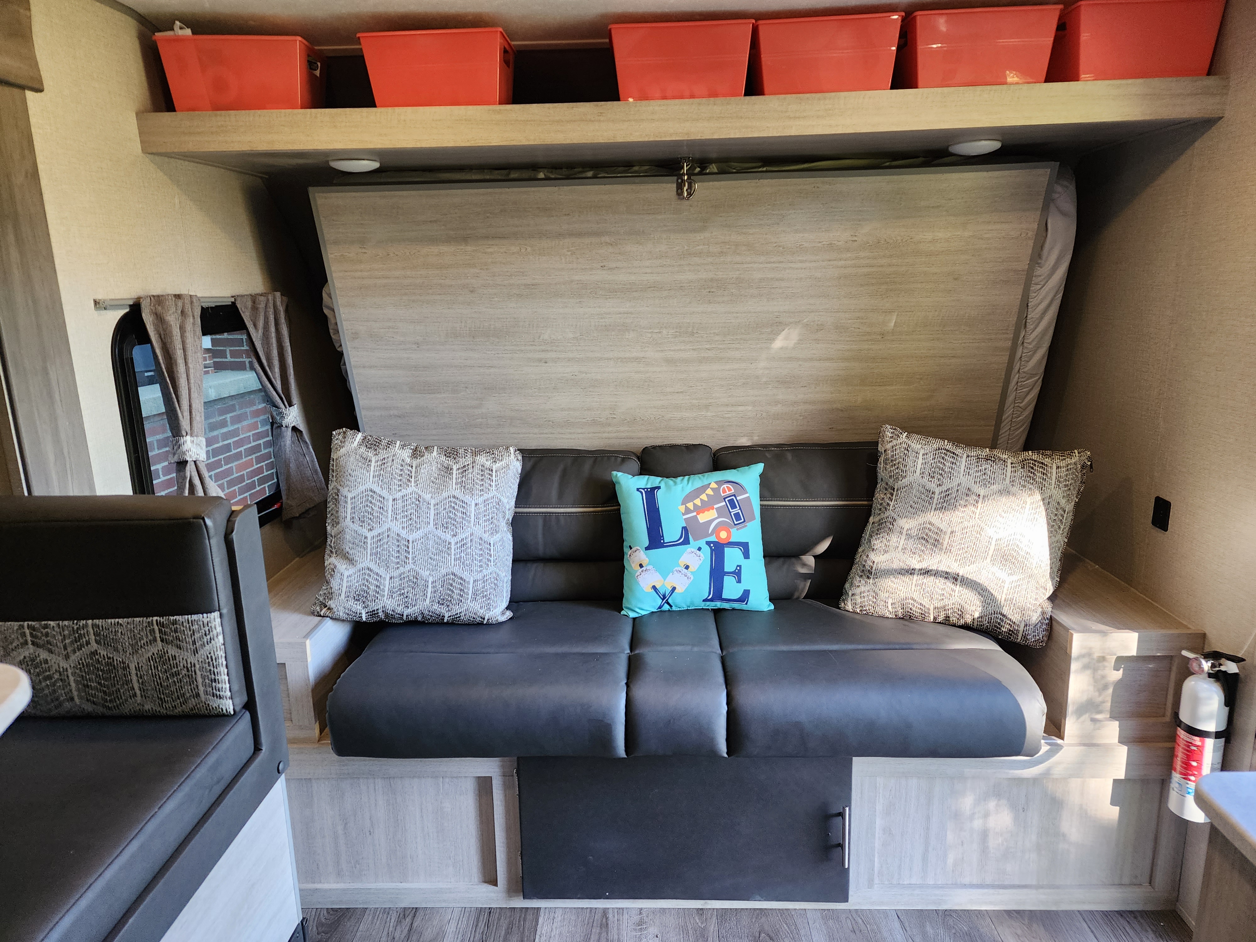 Cozy RV rental interior with a dark couch, decorative pillows, fold-up bed, and overhead storage bins. Fire extinguisher mounted.. Forest River Coachmen Catalina Summit 2022