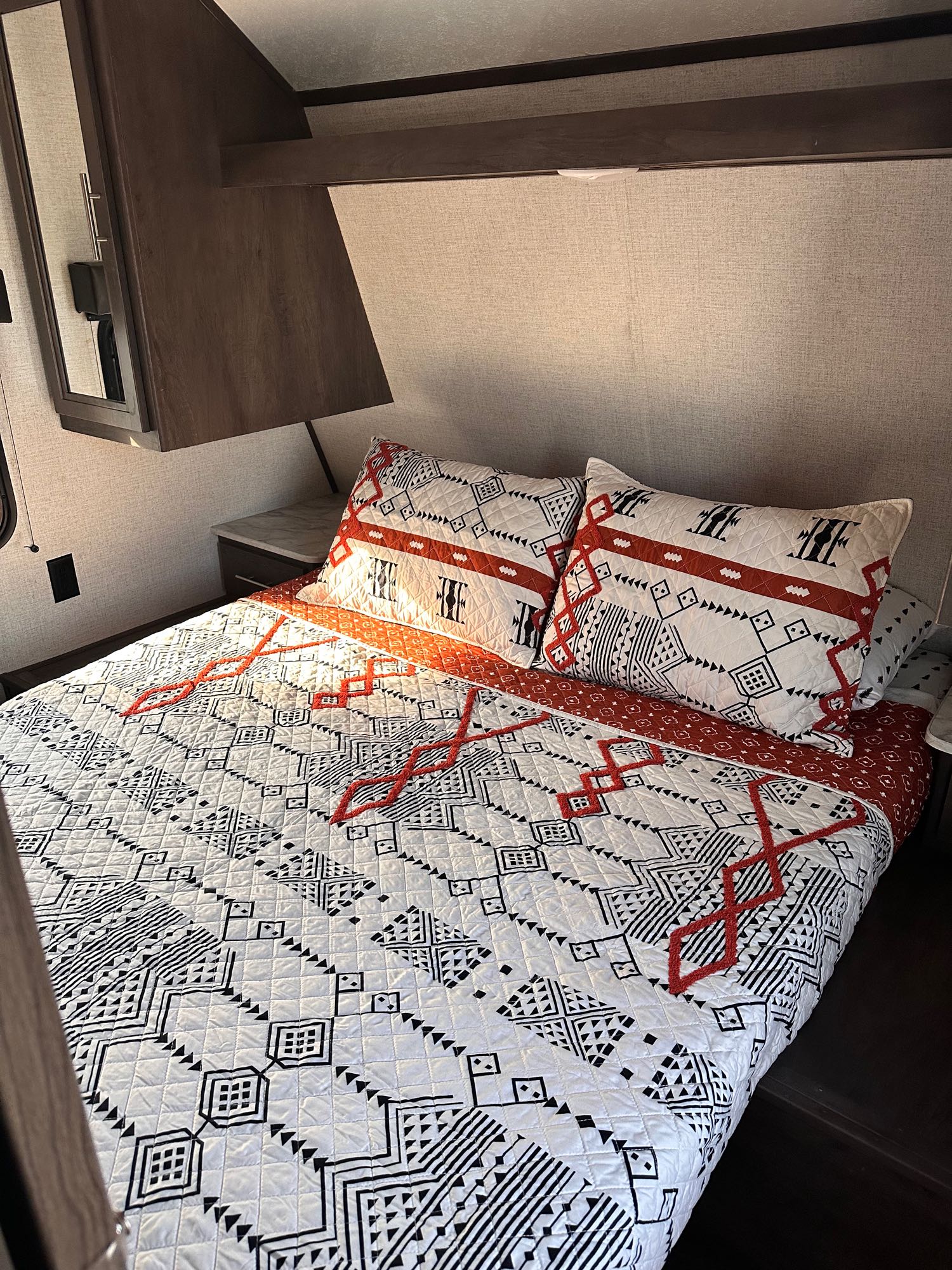 Cozy RV rental bedroom featuring modern white, black, and red geometric bedding, nightstand, and mirrored overhead storage.. Dutchmen Coleman Lantern 2021