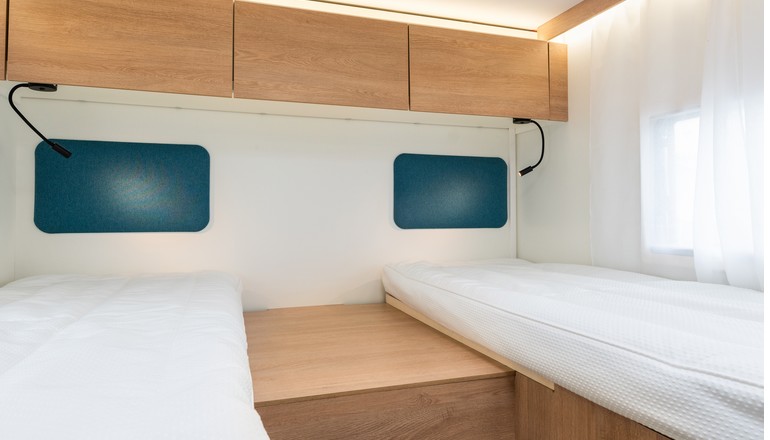 Twin beds in an RV rental bedroom, featuring overhead cabinets, reading lamps, and a window with sheer curtains.. JOA 75T 2025