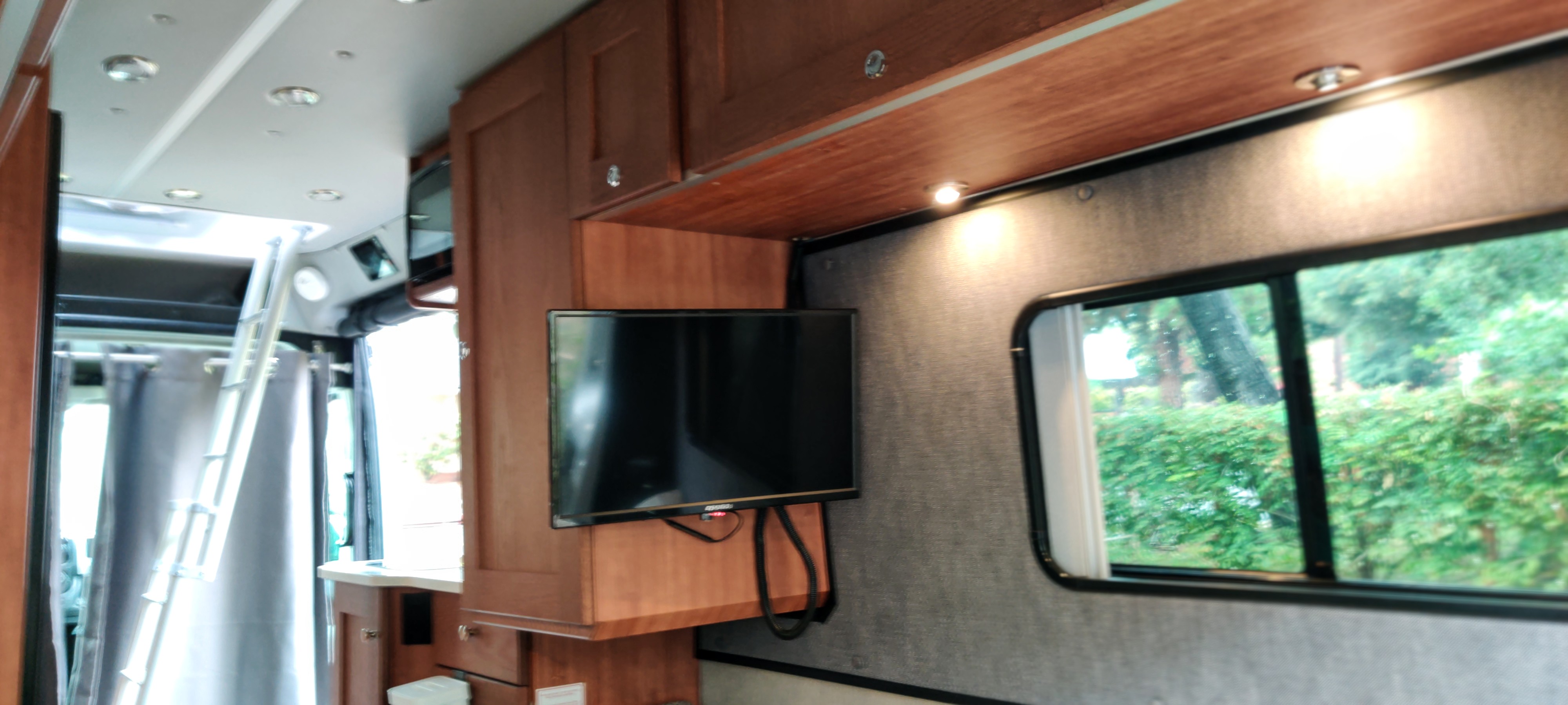 RV rental interior: wooden cabinets, wall-mounted TV, and a ladder to an upper bunk, with a scenic window view.. Roadtrek Roadtrek Class B 2021