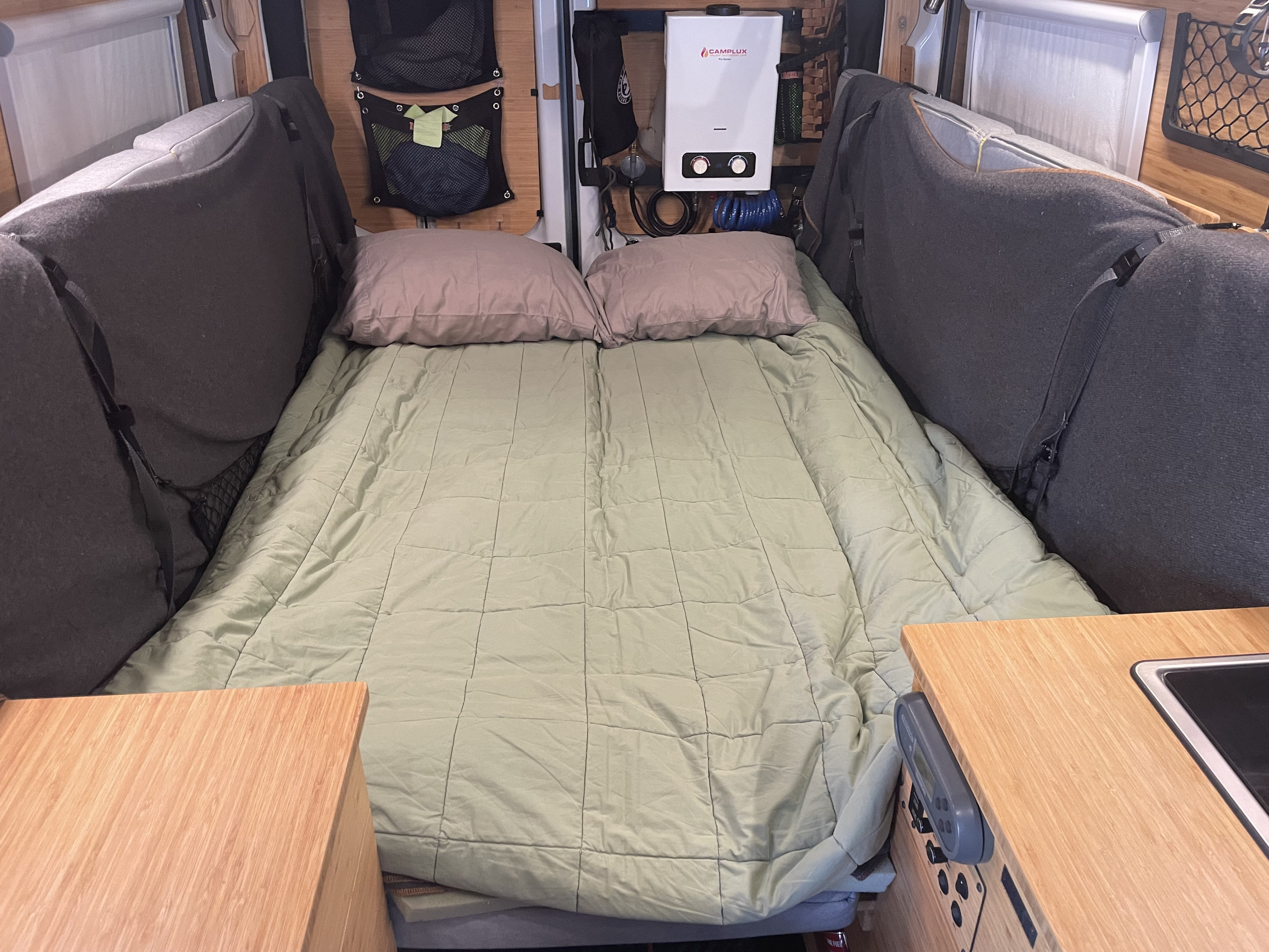 Inside a spacious RV rental: a neatly arranged bed, soft pillows, and essential amenities like a water heater and compact kitchen.. Ford Transit Custom 2019