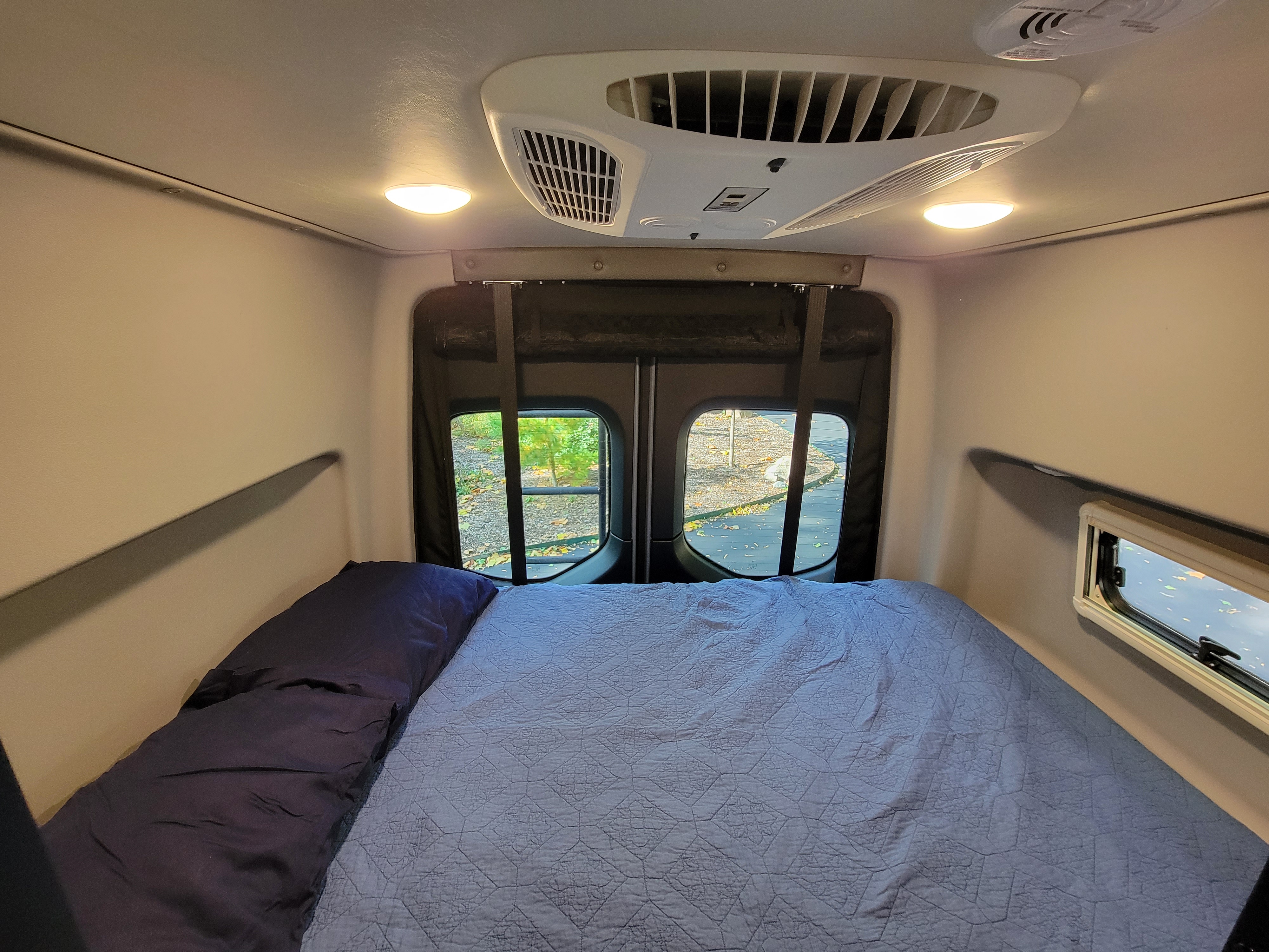 Inside a comfortable RV rental: A cozy bed with blue bedding, rear windows showing nature, and overhead AC unit & lights.. Winnebago Revel 2022