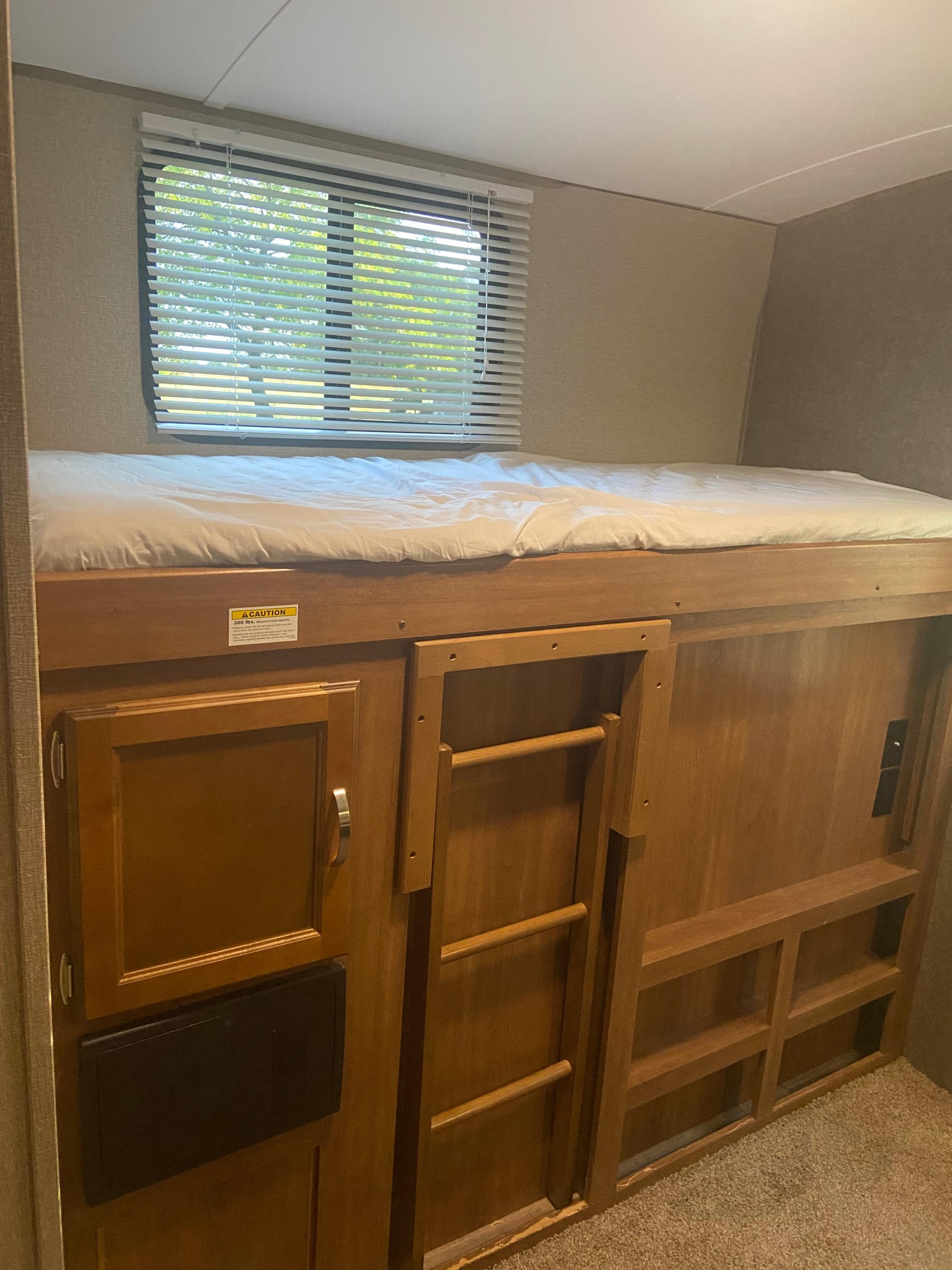 An RV rental features a top bunk bed with white bedding, a window with blinds, a ladder, and built-in storage shelves below.. Keystone RV Springdale 2018