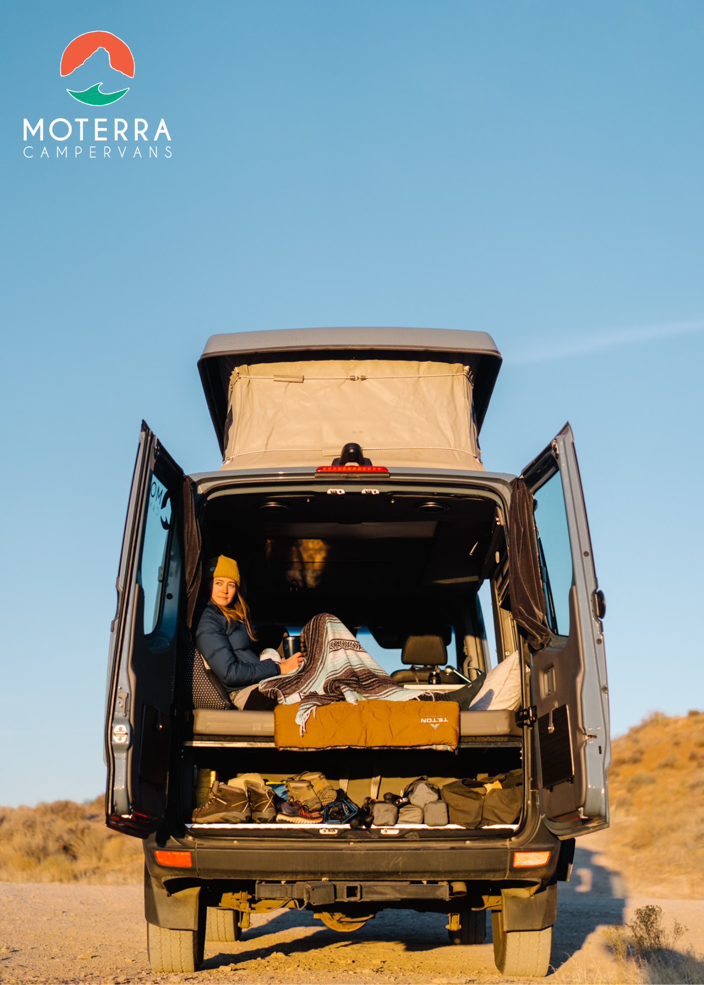 Woman relaxes in a gray RV rental campervan. Rear doors open, pop-top up, showing a desert view and gear inside.. Mercedes-Benz Sprinter 2021