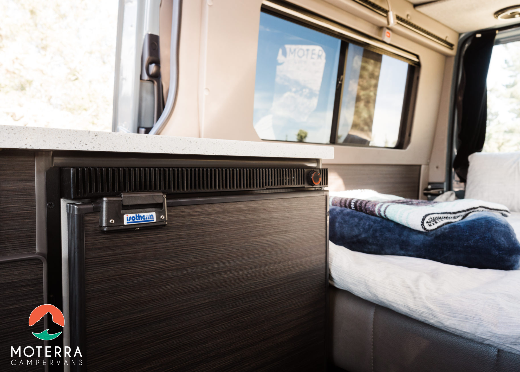 Inside an RV rental: a dark wood-look Isotherm fridge beneath a speckled countertop, next to a cozy bed. Ideal campervan comfort.. Mercedes-Benz Sprinter 2021