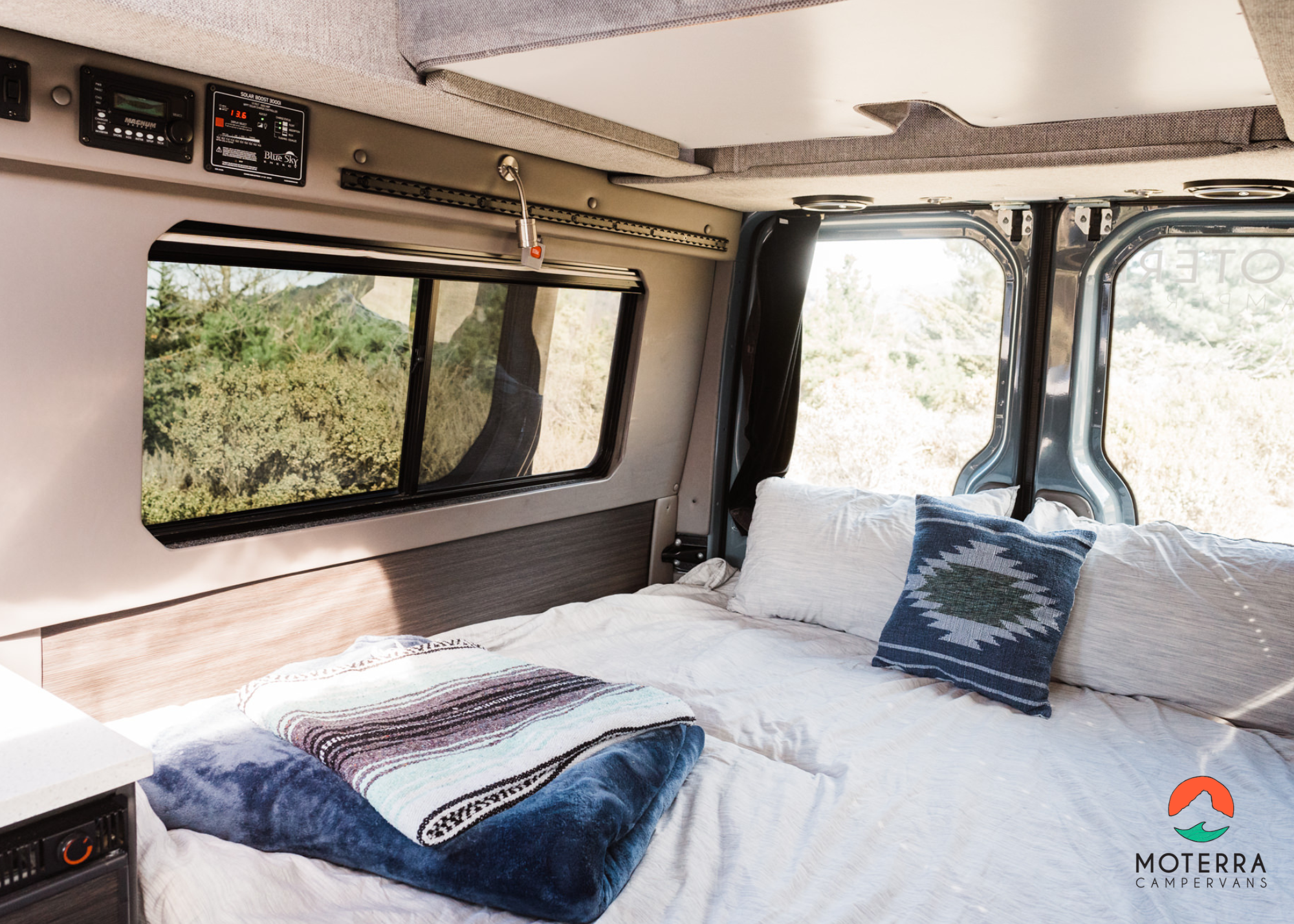 Inside a comfy RV rental: a spacious bed with pillows & blankets, control panels, and a scenic window view.. Mercedes-Benz Sprinter 2020