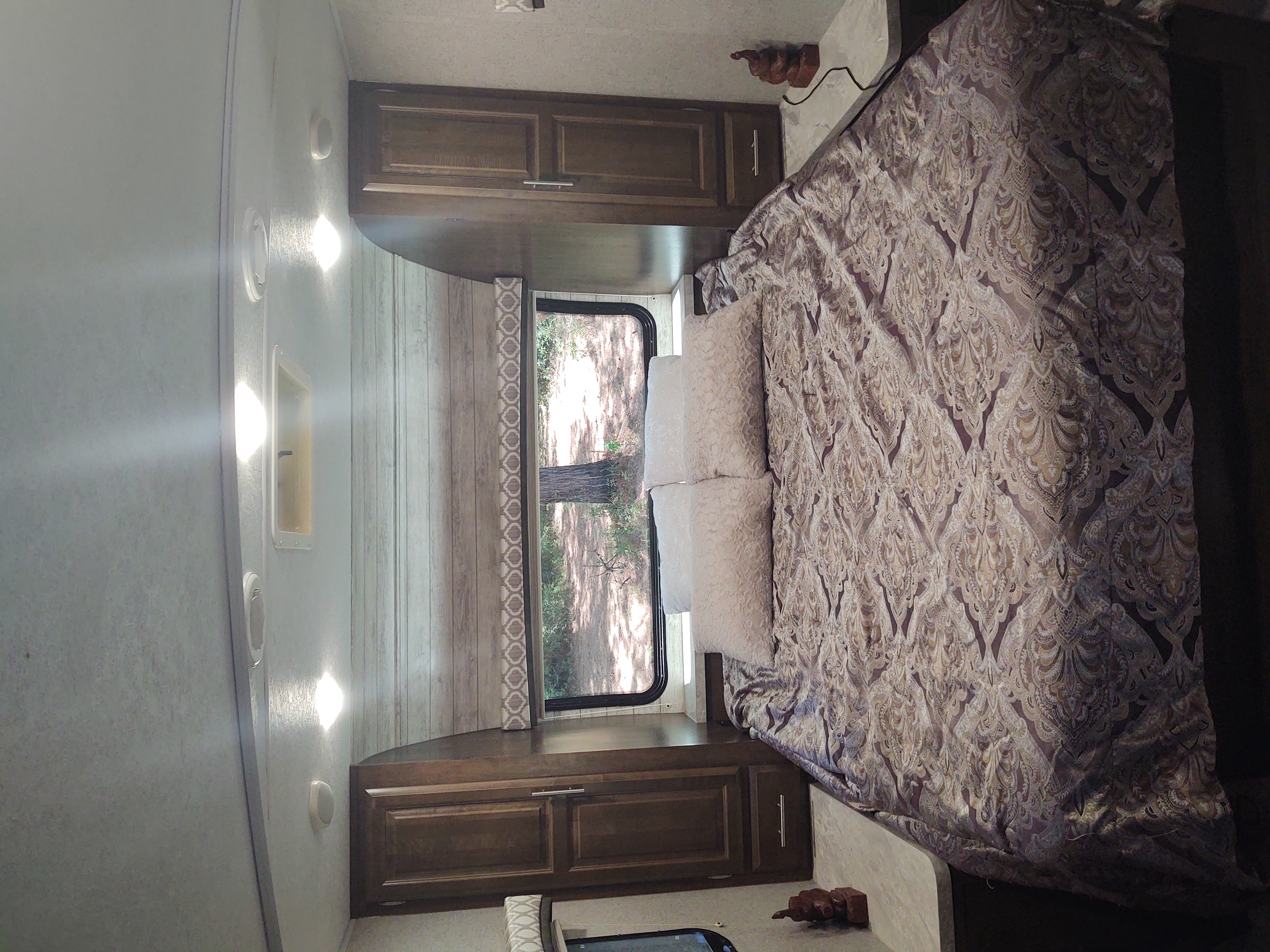 Inside an RV rental bedroom: a comfy bed with patterned linens, overhead storage, and a scenic window view.. Forest River Flagstaff Micro Lite 2019