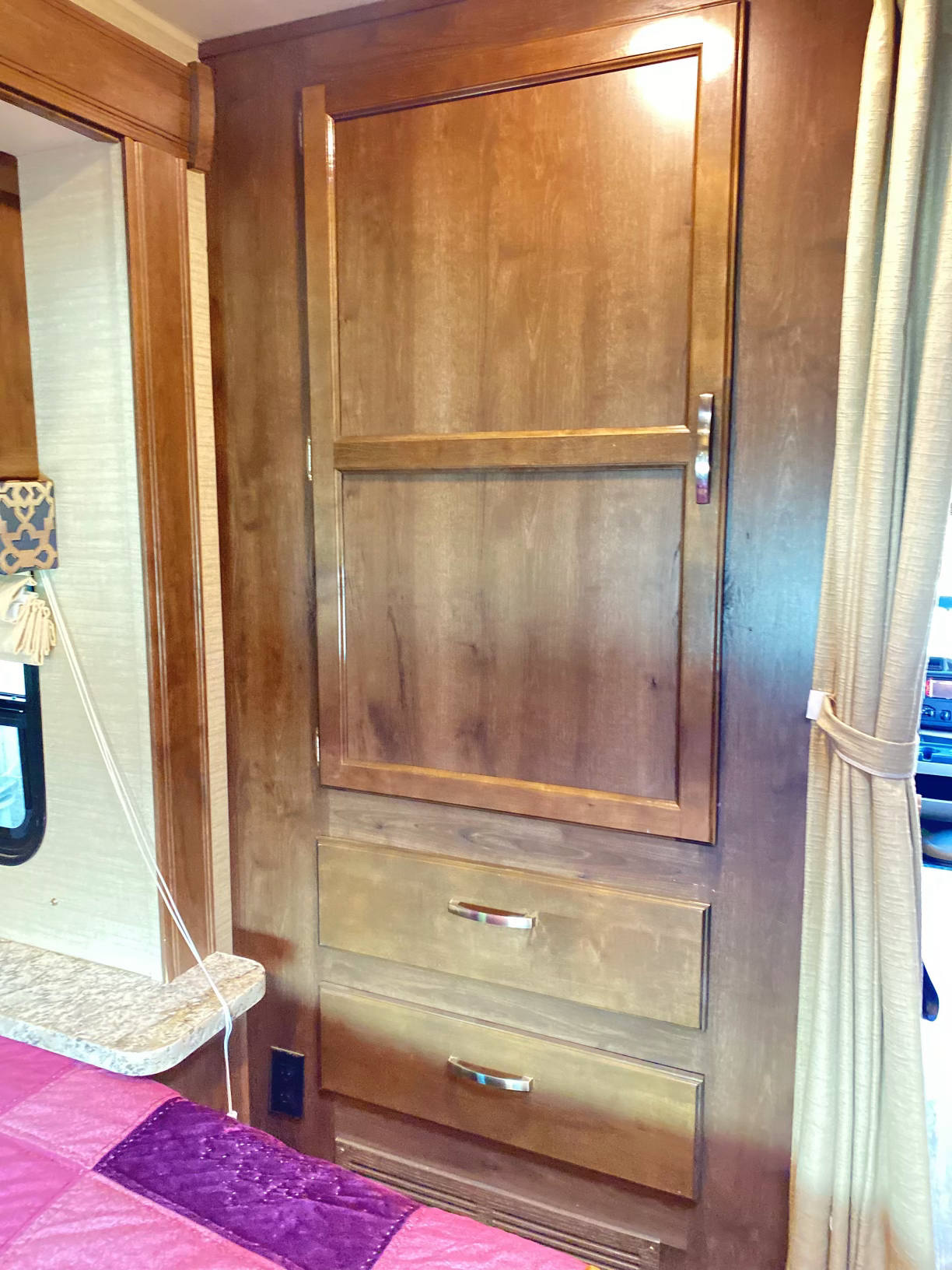 RV rental bedroom features a wood cabinet with a door and two drawers, a tied-back curtain, and a bed.. Jayco Redhawk 2018