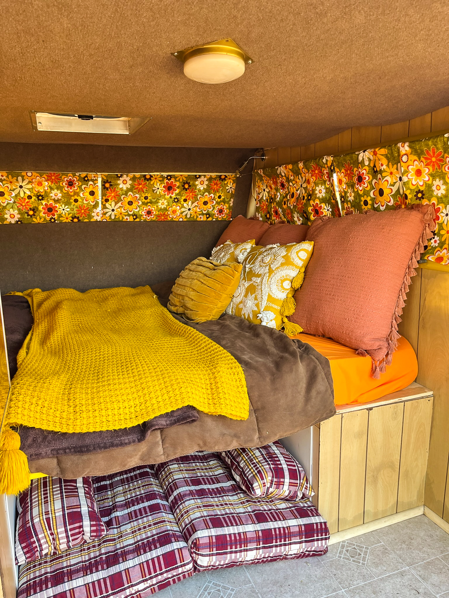 Cozy, retro RV rental interior featuring vibrant floral curtains, yellow knit blanket, plush pillows, and wood paneling.. Custom Camper Trailer 1987