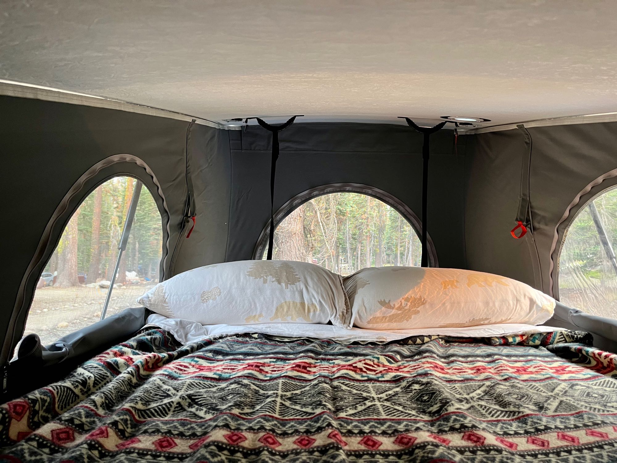 Cozy interior of an RV rental rooftop tent with a comfy bed, patterned blanket, and nature-themed pillows, overlooking a forest.. Winnebago Solis 2021