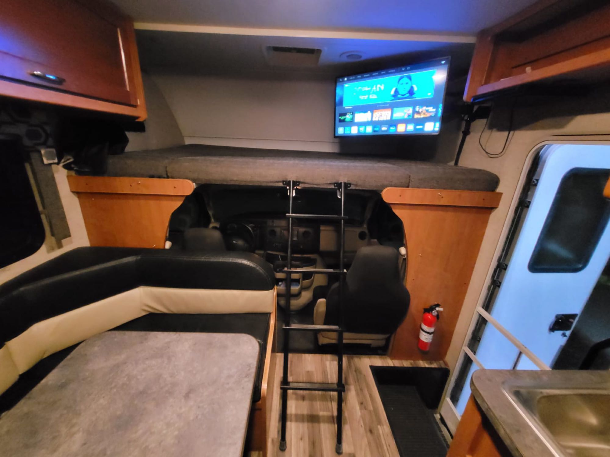 An RV rental's interior, showing a front bunk bed with ladder, mounted TV, dining area, and driver's cabin.. Winnebago Minnie Winnie 2018