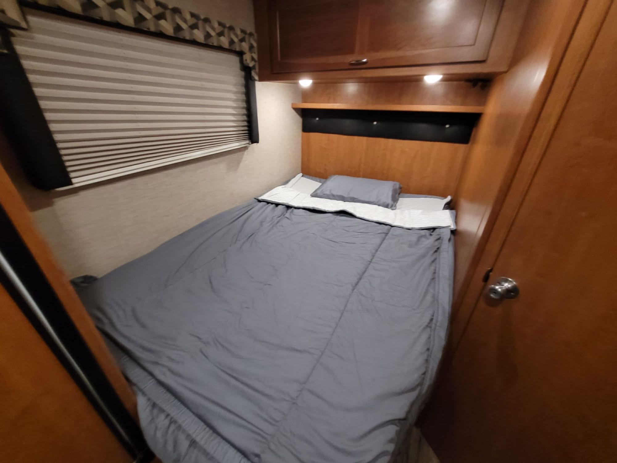 Compact RV rental sleeping area with a gray bed, overhead wood cabinets, and pleated window blinds. Features built-in storage and reading lights.. Winnebago Minnie Winnie 2018