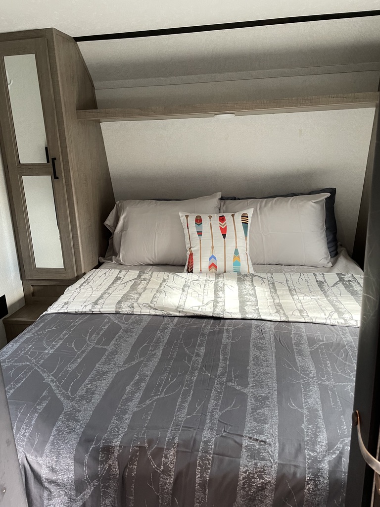Inside an RV rental: A comfortable bedroom with a gray tree-patterned bedspread and colorful paddle-design pillow.. Keystone RV Hideout 2020