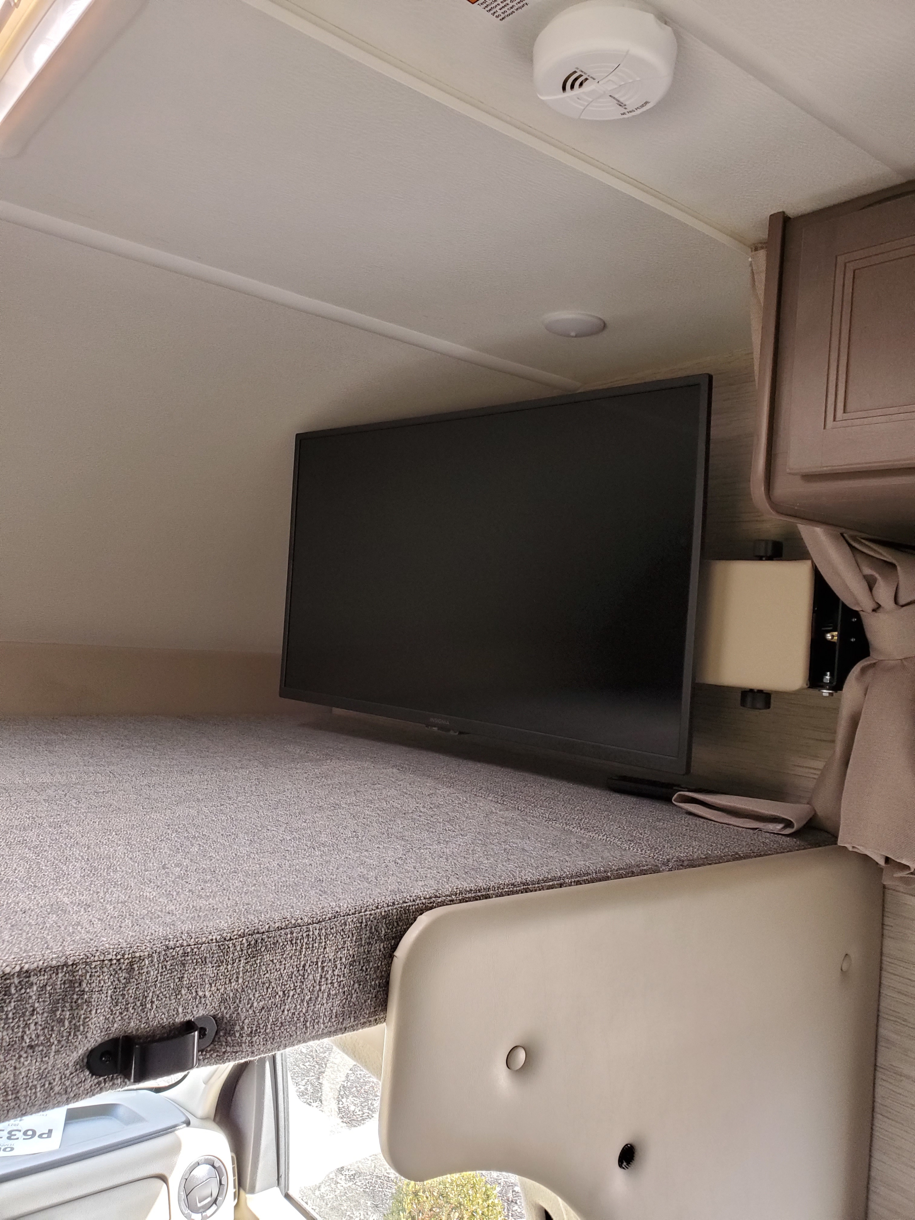 An RV rental's over-cab bunk area with a mounted Insignia TV, showing safety features like a smoke detector.. Thor Coleman 2023