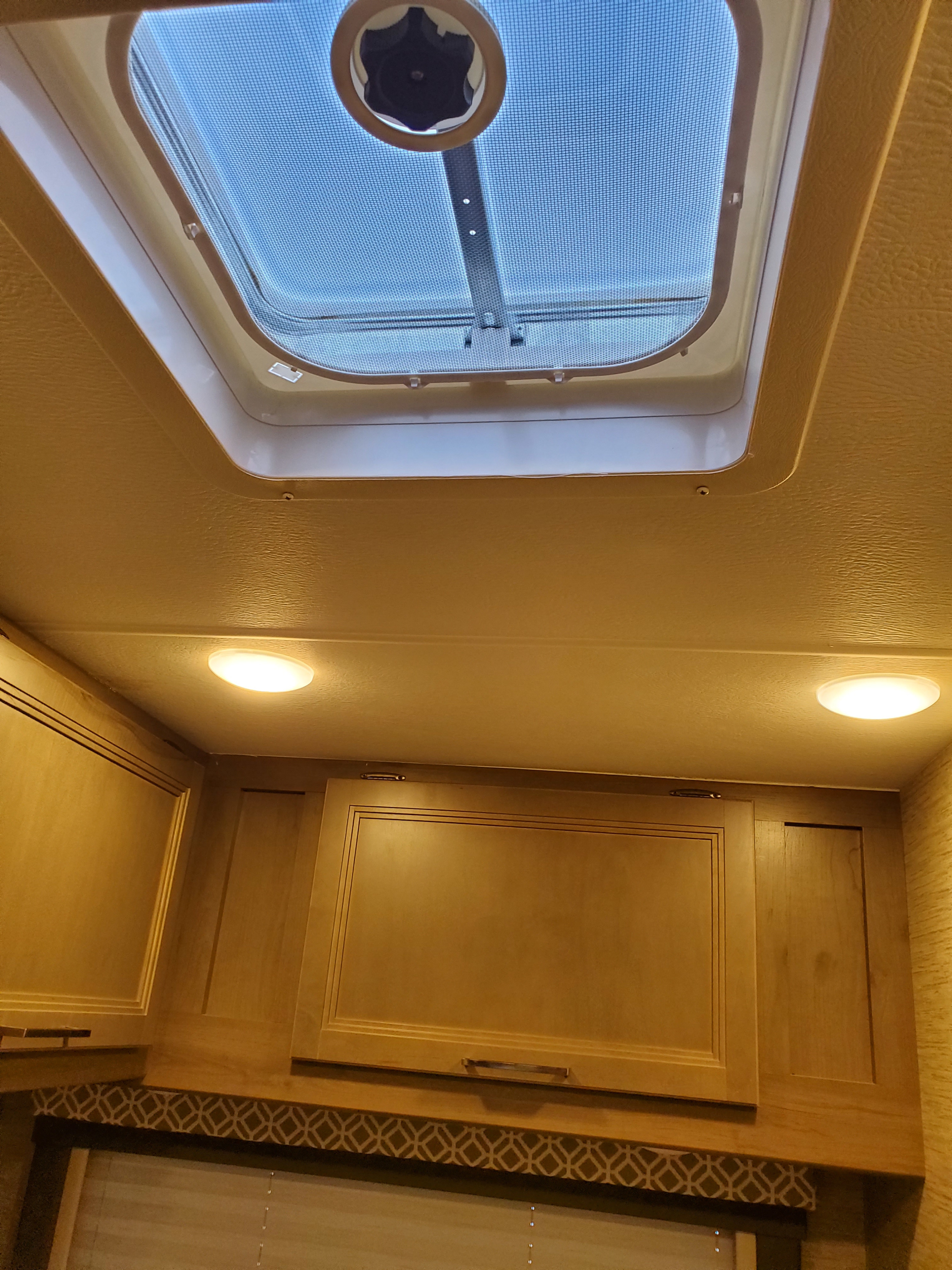 An RV rental interior view showing a large ceiling skylight with a screen and fan, two bright lights, and wooden cabinets.. Thor Coleman 2023