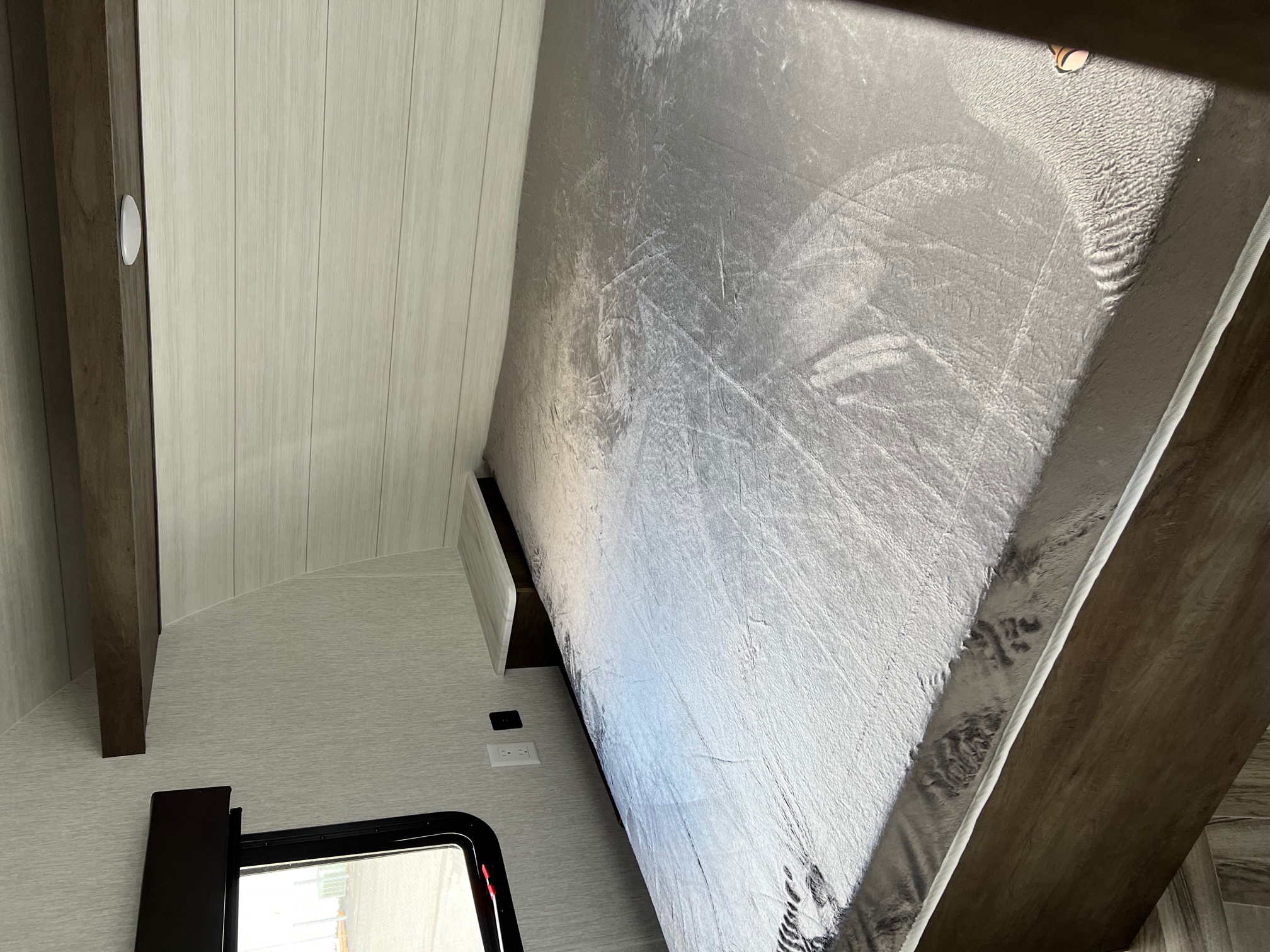 RV rental interior: A compact bedroom featuring a textured bed, light paneling, and wall outlets.. Forest River Wildwood X-Lite 2023