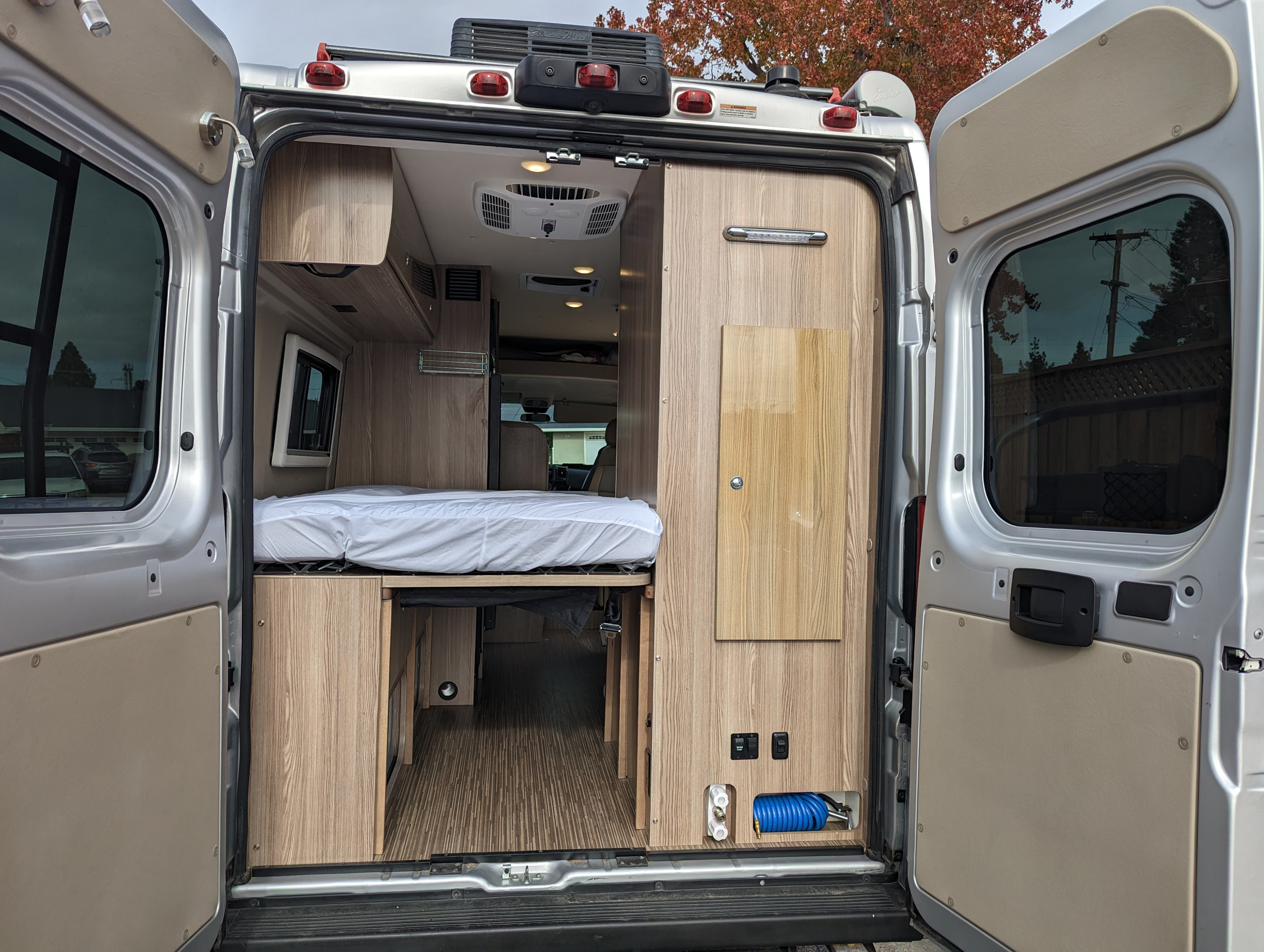 Rear view of an rv rental's interior, featuring a bed, under-bed storage, and a utility panel with hose, in a modern camper van.. Winnebago Travato 2020