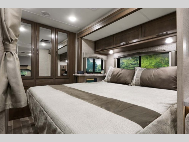 Spacious RV rental bedroom with a cozy bed, ample storage, and large windows revealing a lush green view.. Thor Chateau 2023