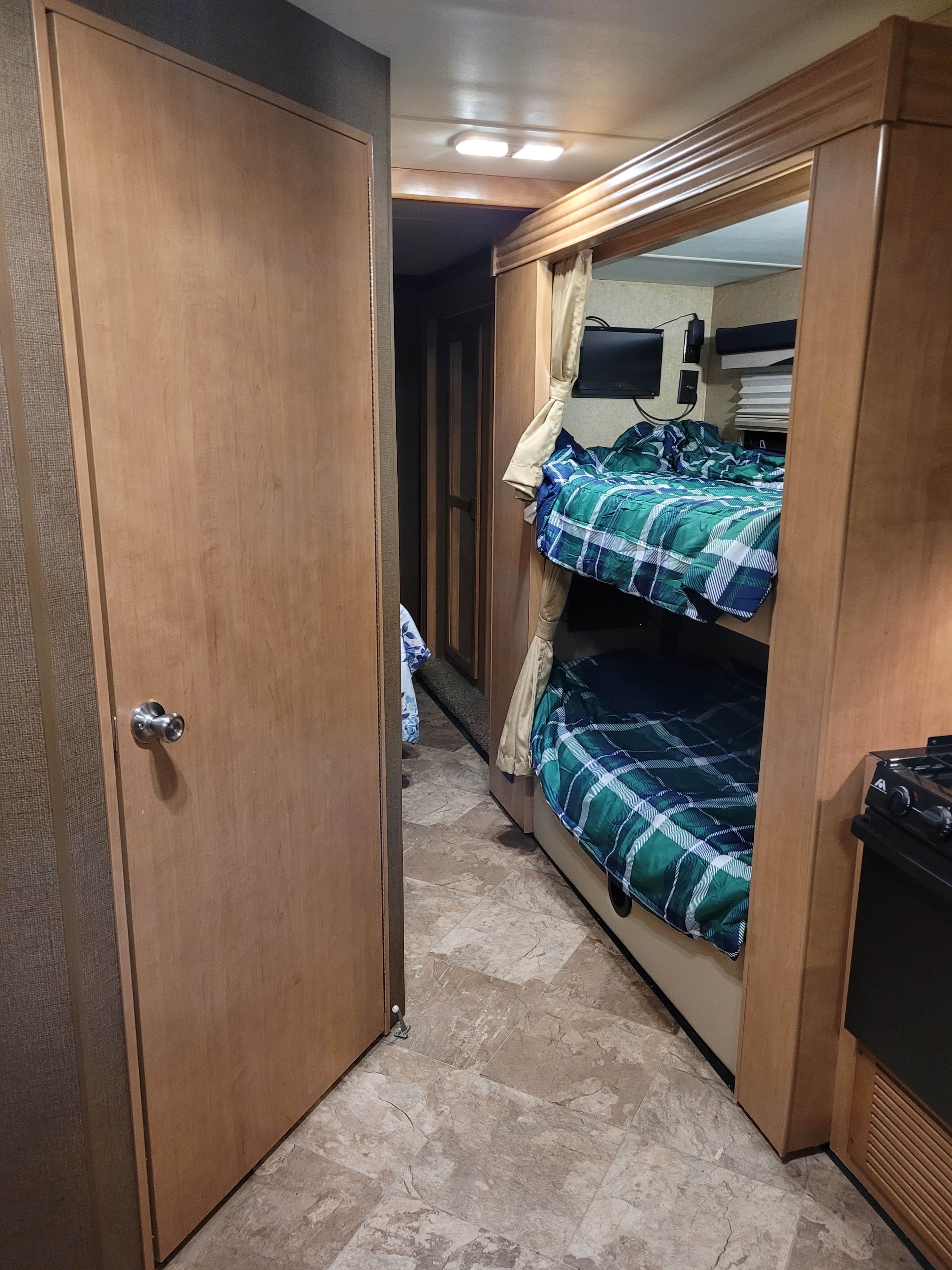 RV rental interior features bunk beds with plaid bedding, a wall-mounted TV, and a partial kitchen view on patterned flooring.. Thor A.C.E. 2017