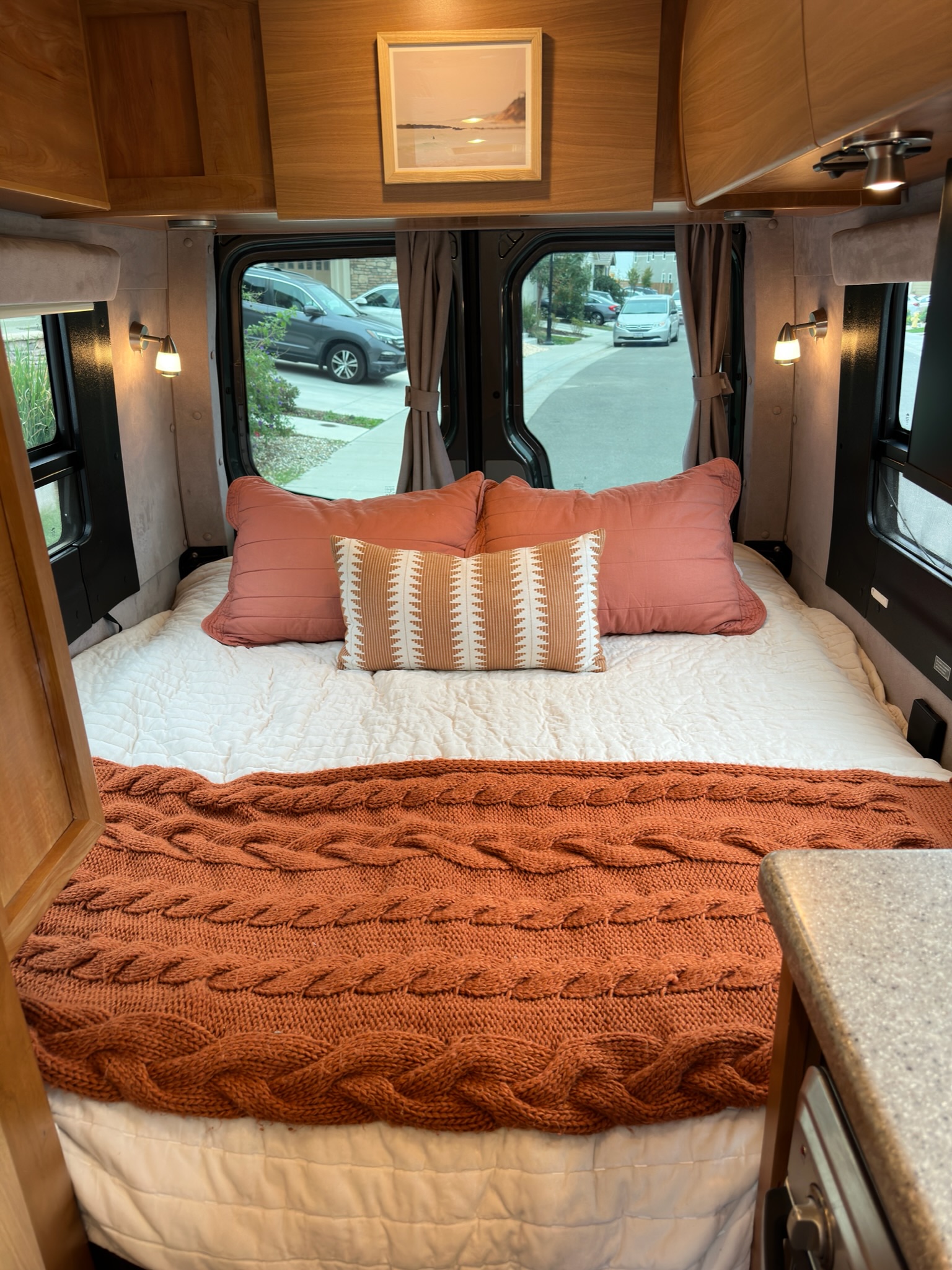 Cozy RV rental interior featuring a comfy bed with a warm cable knit blanket, pillows, wooden cabinets, and outside views.. ERA Era 2010