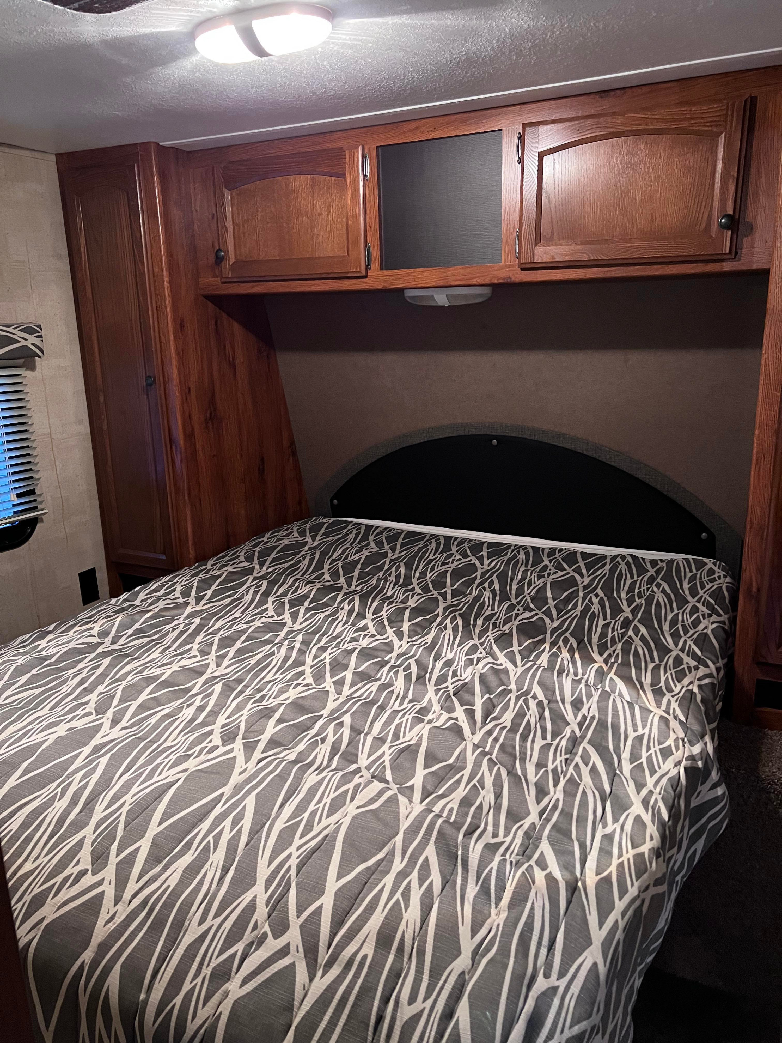 RV rental bedroom: A bed with a gray and white abstract patterned comforter, dark headboard, and wood overhead cabinets.. Keystone RV Hideout 2015