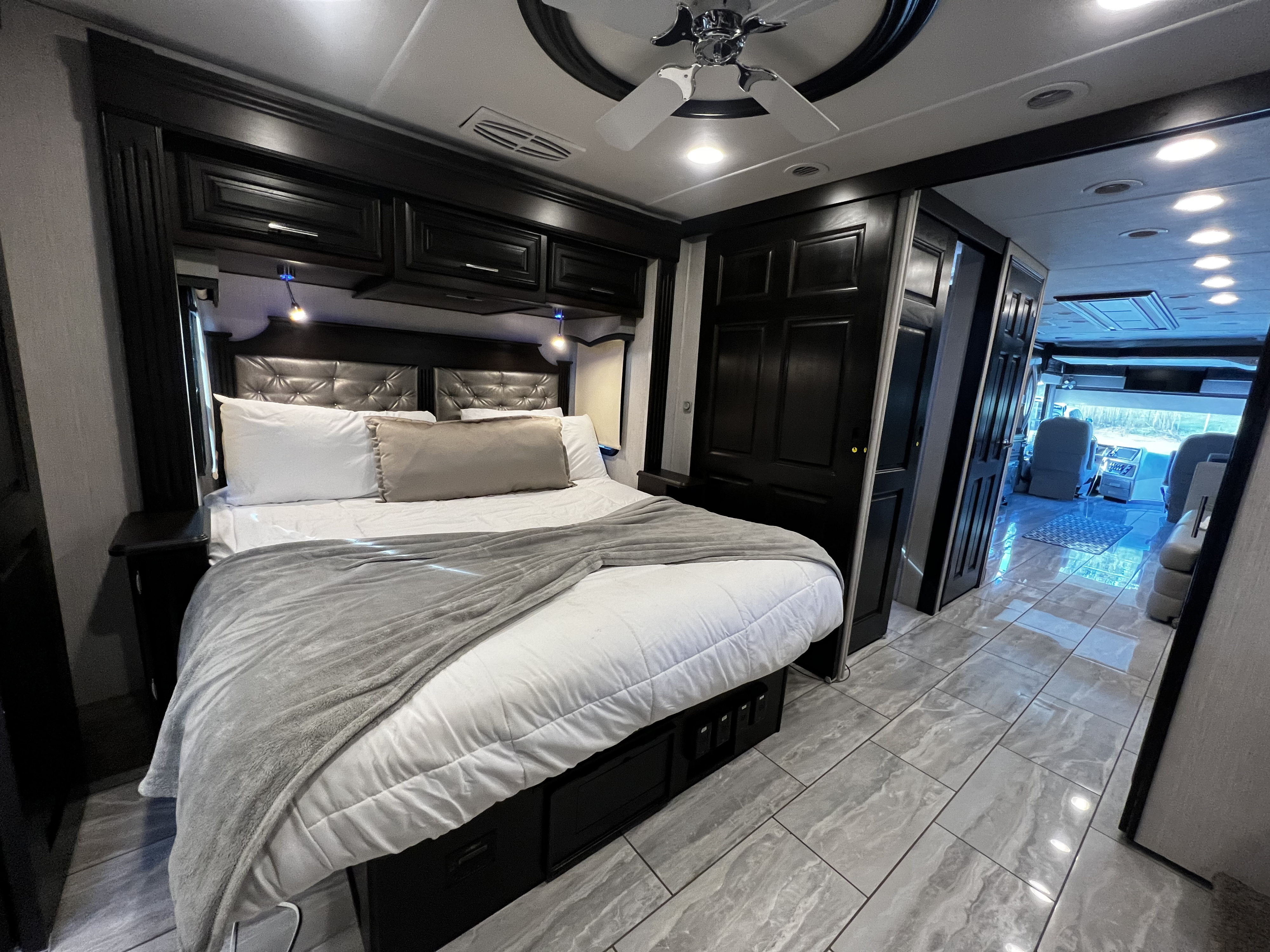 Luxurious RV rental bedroom featuring a large bed, dark wood decor, modern tiled floor, and a glimpse of the cockpit area.. Forest River Berkshire 2022