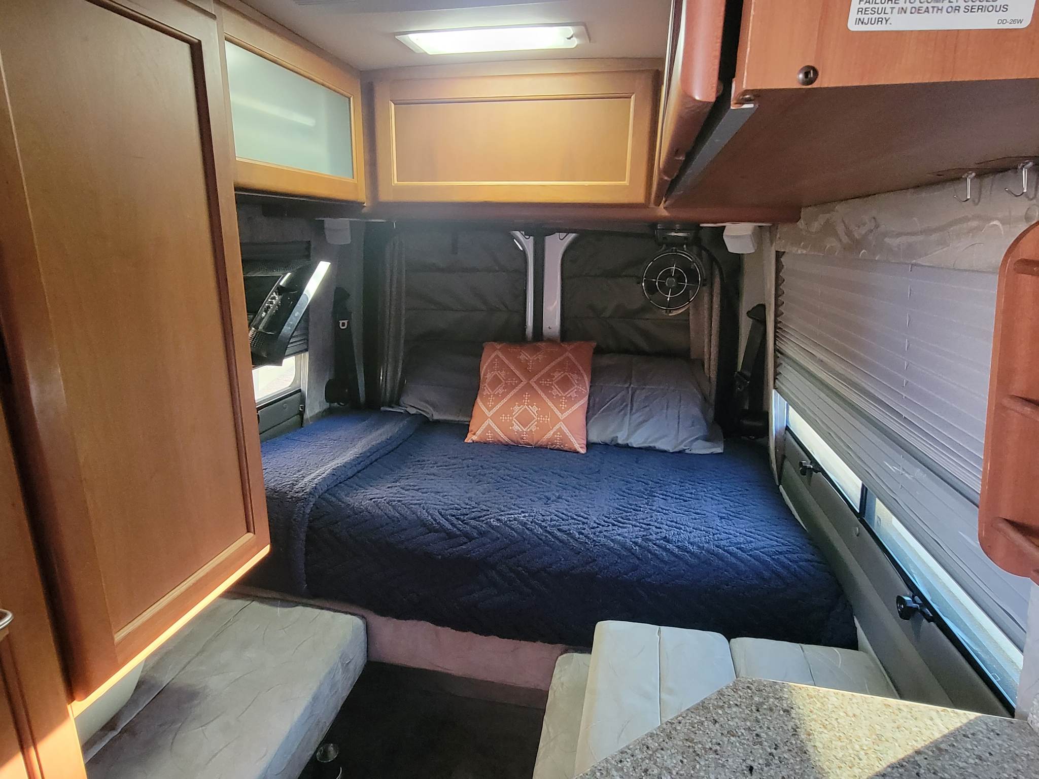 RV rental bedroom: a bed with blue comforter & orange pillow, overhead wood cabinets, fan, & blinds.. Roadtrek Roadtrek Class B 2012