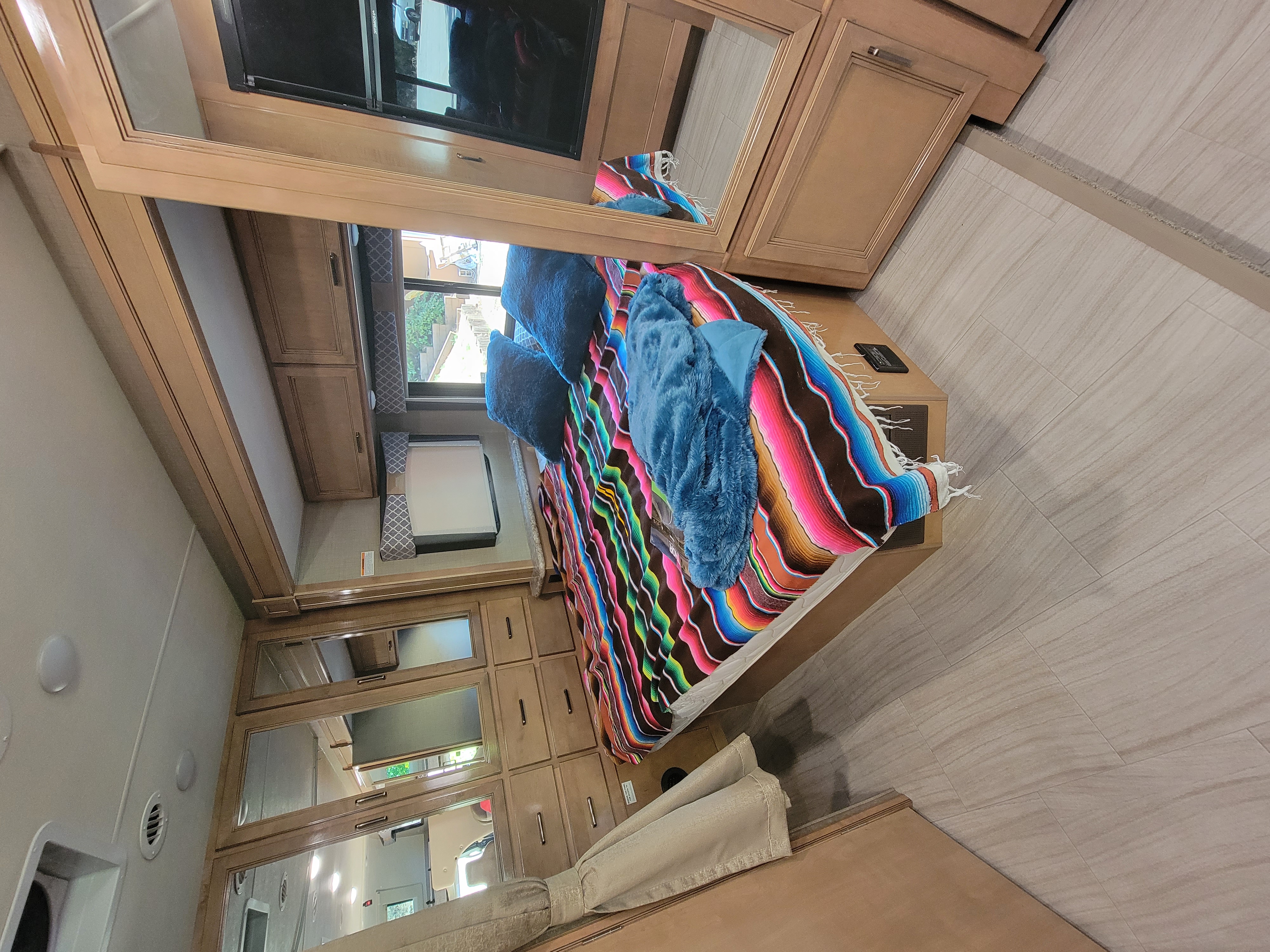Cozy RV rental bedroom with a vibrant serape blanket, plush blue bedding, built-in wood cabinetry, TV, and light tile flooring.. Thor Quantum 2020