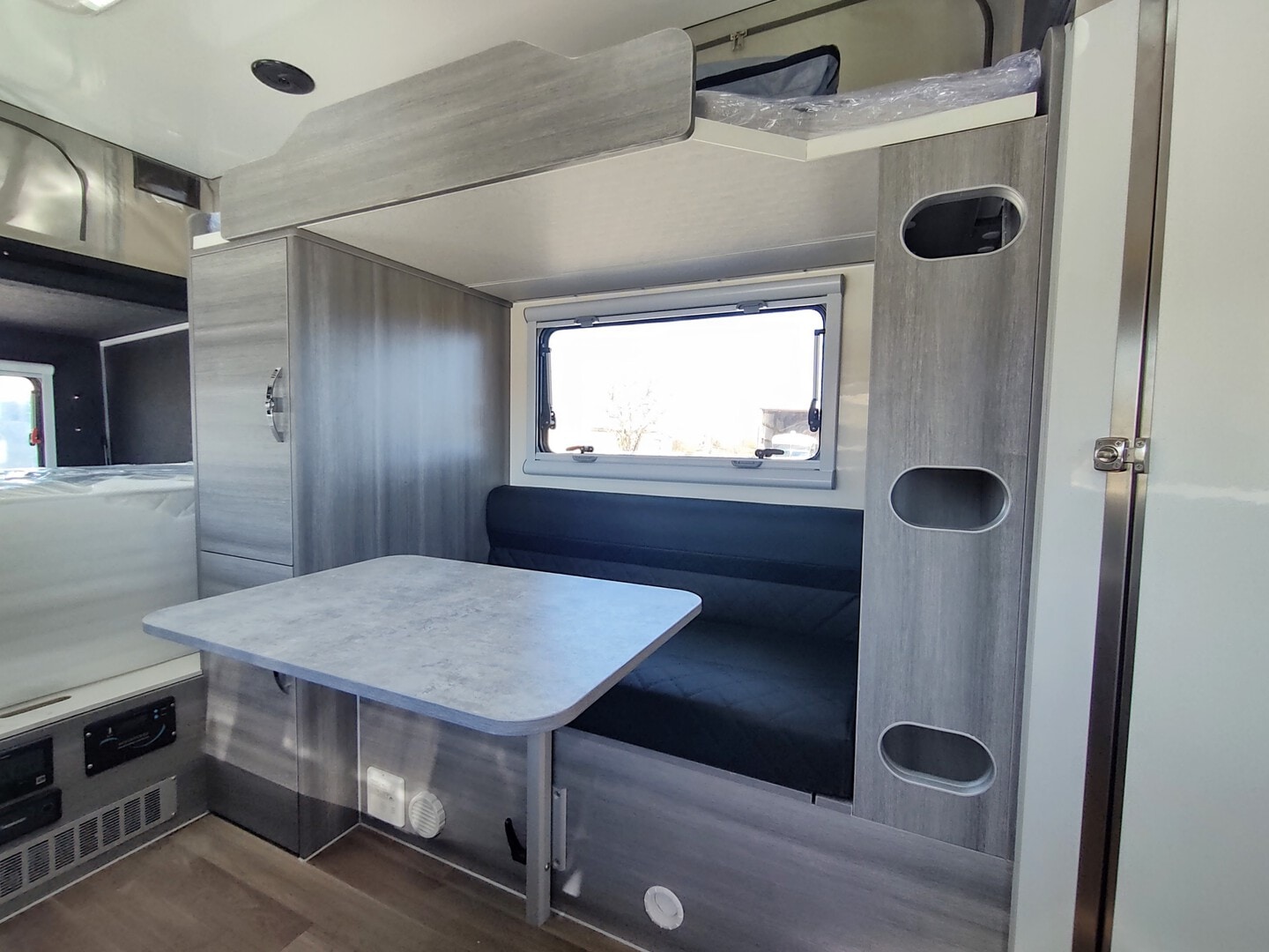 Bright RV rental interior with a gray dinette area, overhead bunk bed, and integrated storage cabinets. A compact, functional living space.. OBI DWELLER 15' 2022