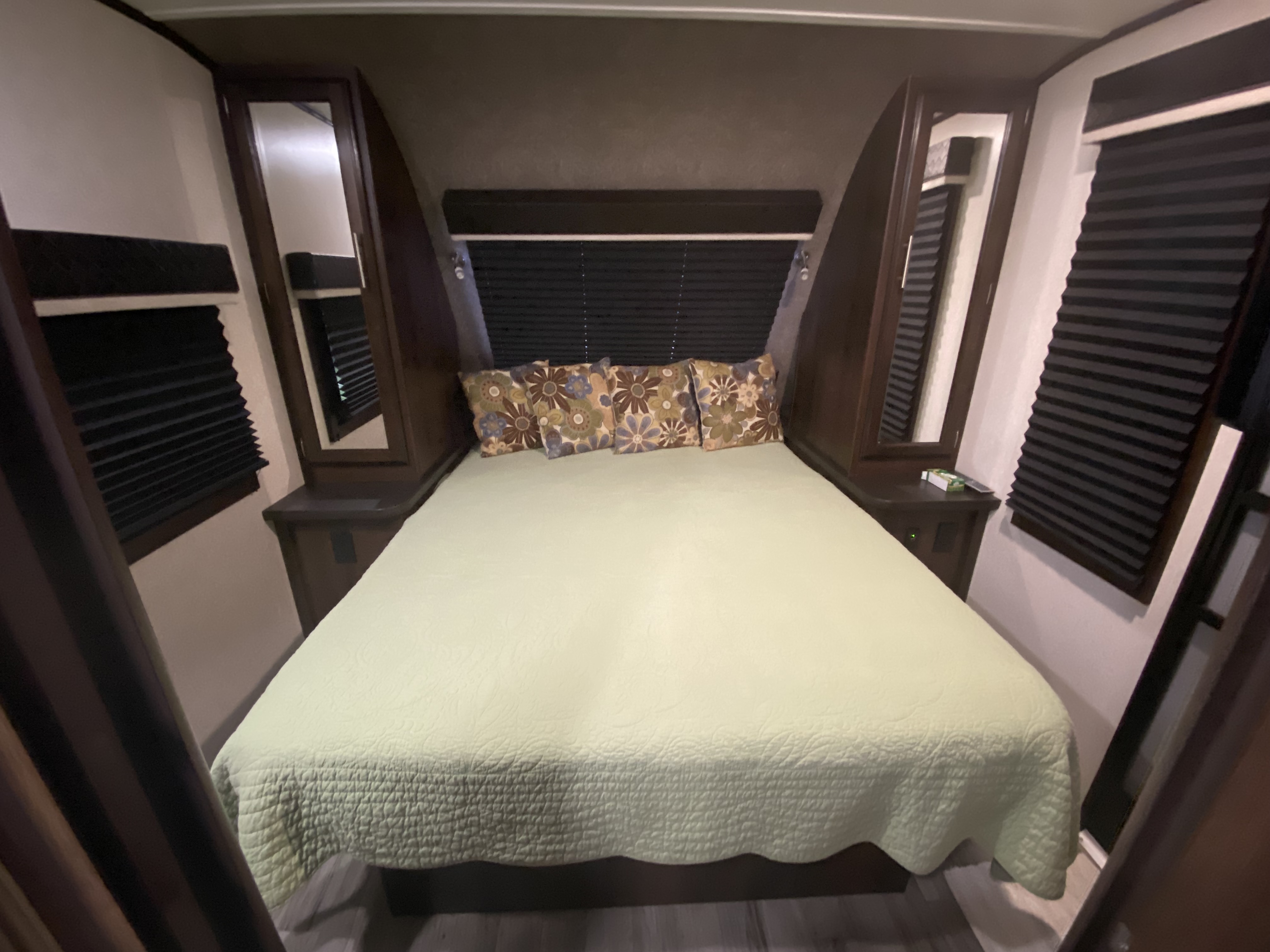 RV rental bedroom featuring a queen bed with light green bedding, patterned pillows, built-in storage, and dark blinds.. Jayco White Hawk 2019