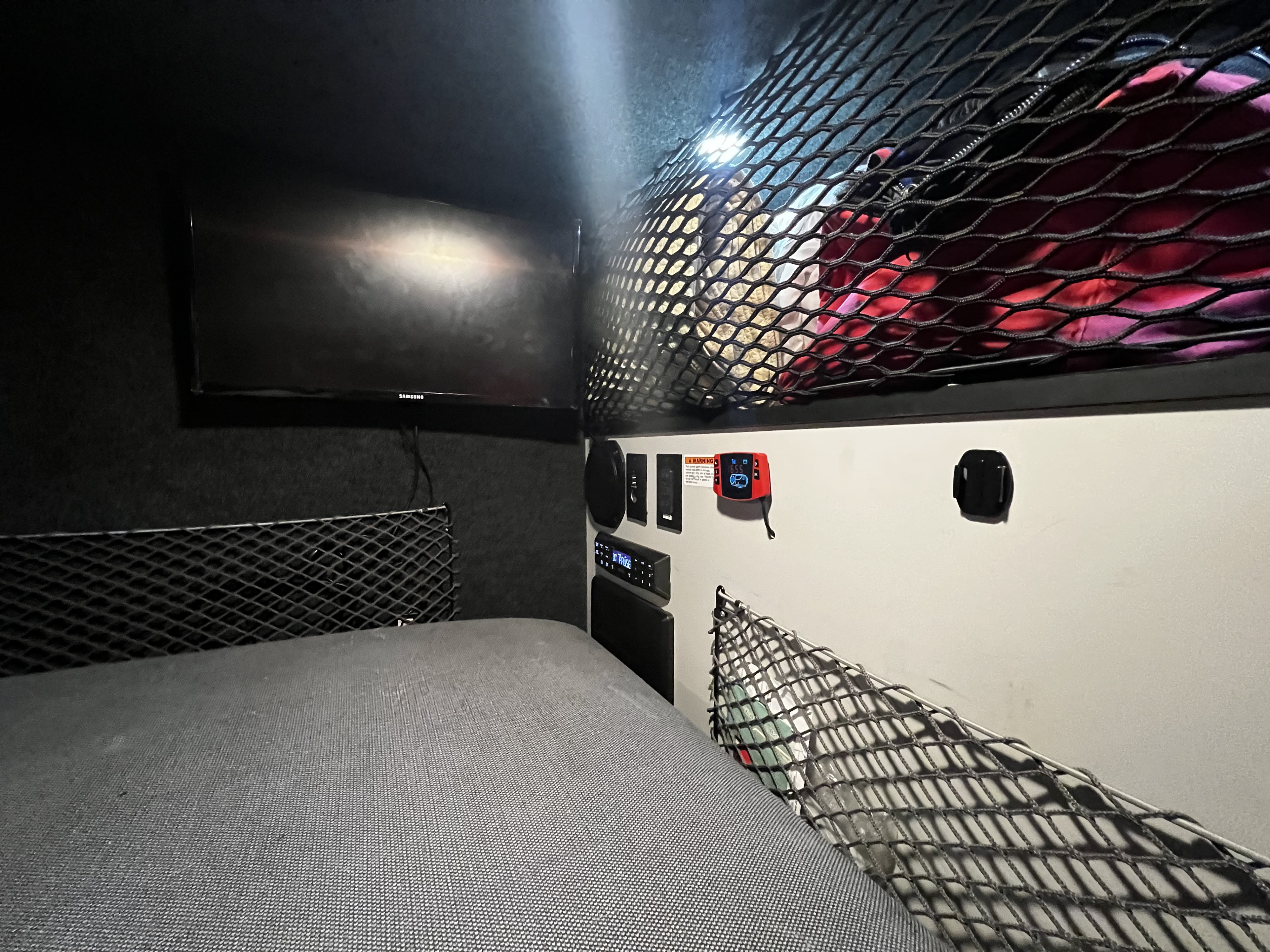 Inside an RV rental: a compact bedroom with a bed, TV, and storage nets overhead and along walls.. Custom Camper 2021