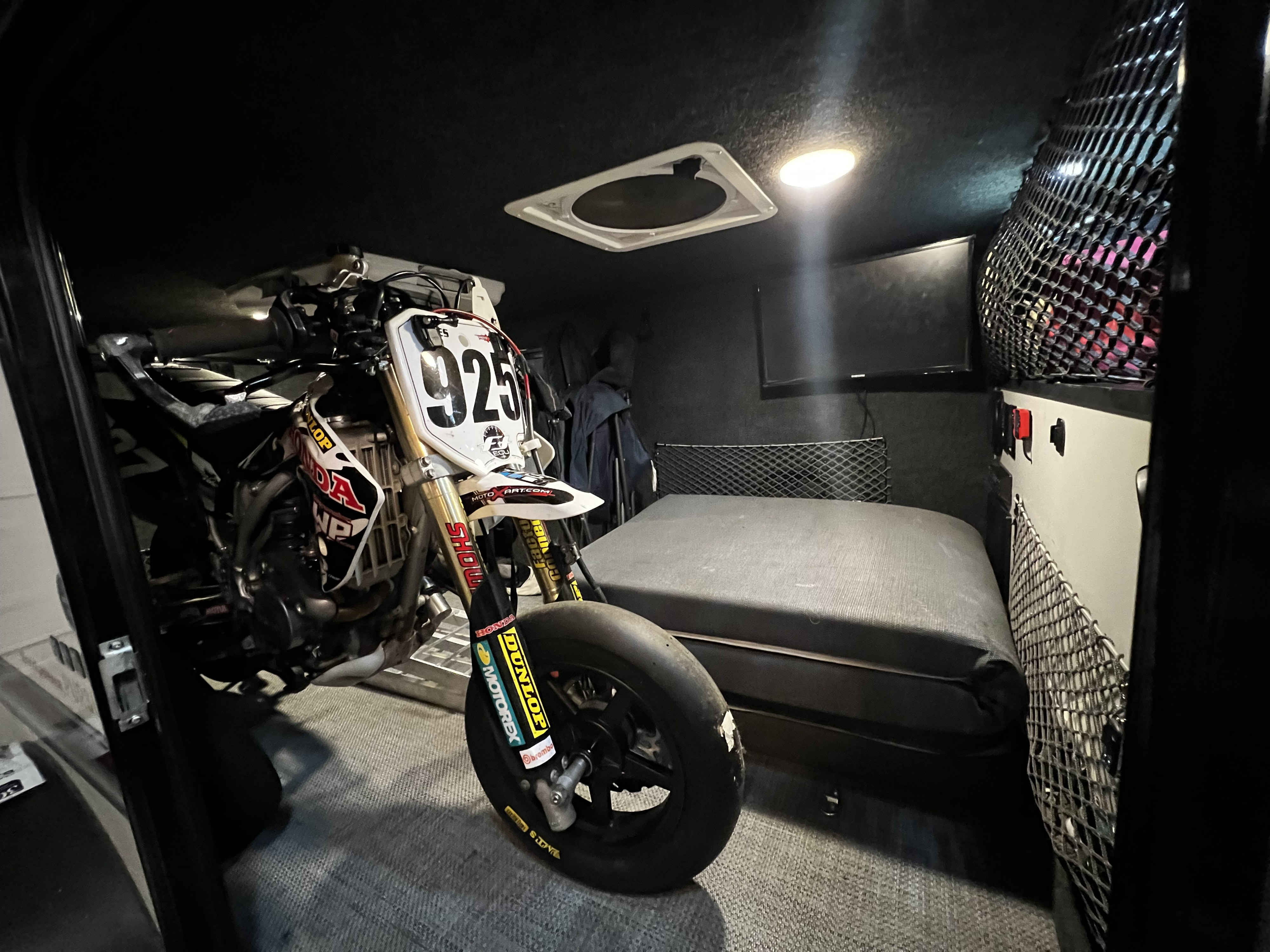 Inside a custom **rv rental**: a dirt bike (925) parked next to a cozy bed, with a TV & ceiling fan.. Custom Camper 2021