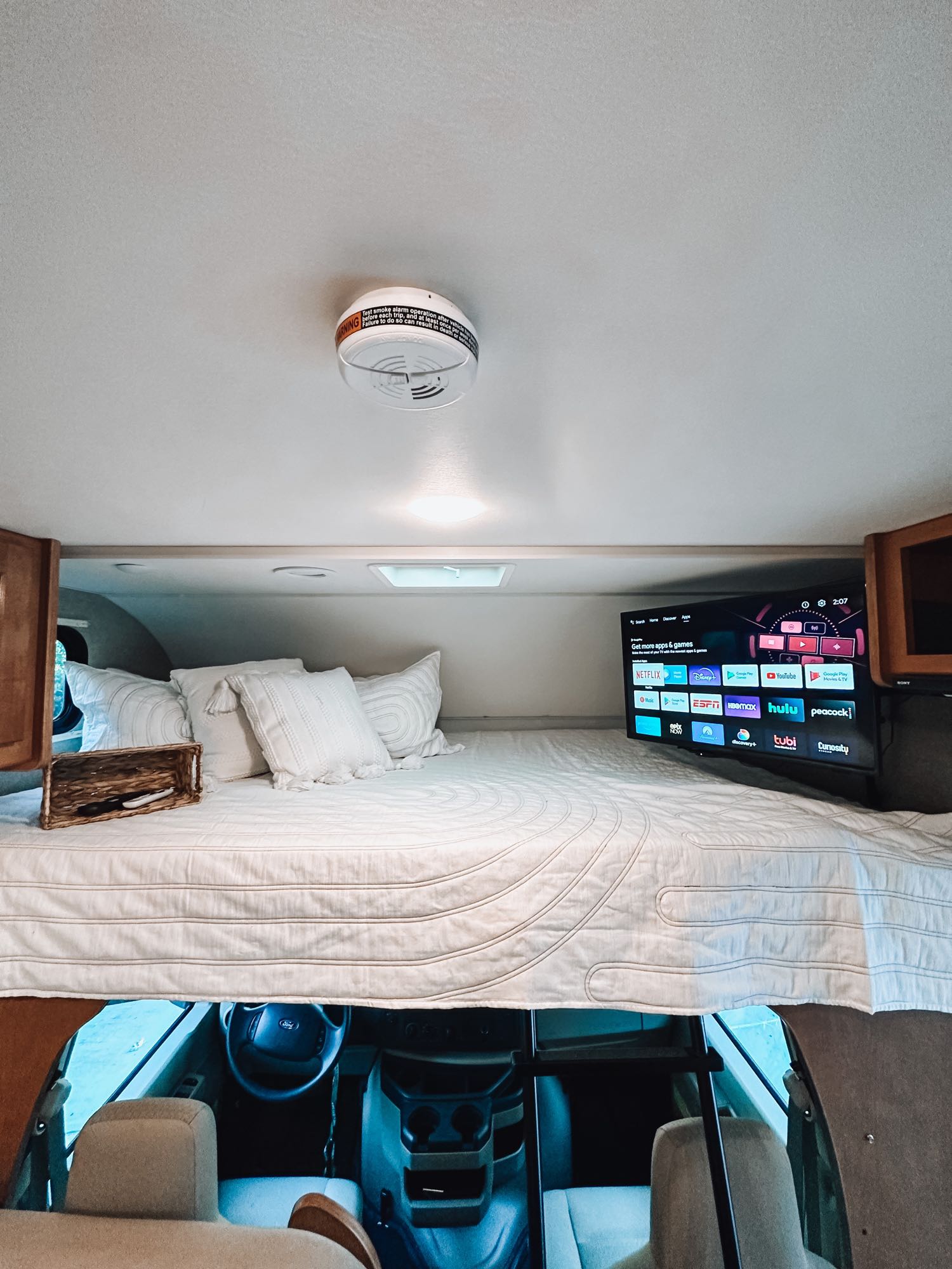 Cozy over-cab bunk bed in an RV rental, featuring a smart TV with streaming apps, soft bedding, and a smoke detector.. Winnebago Spirit 2019