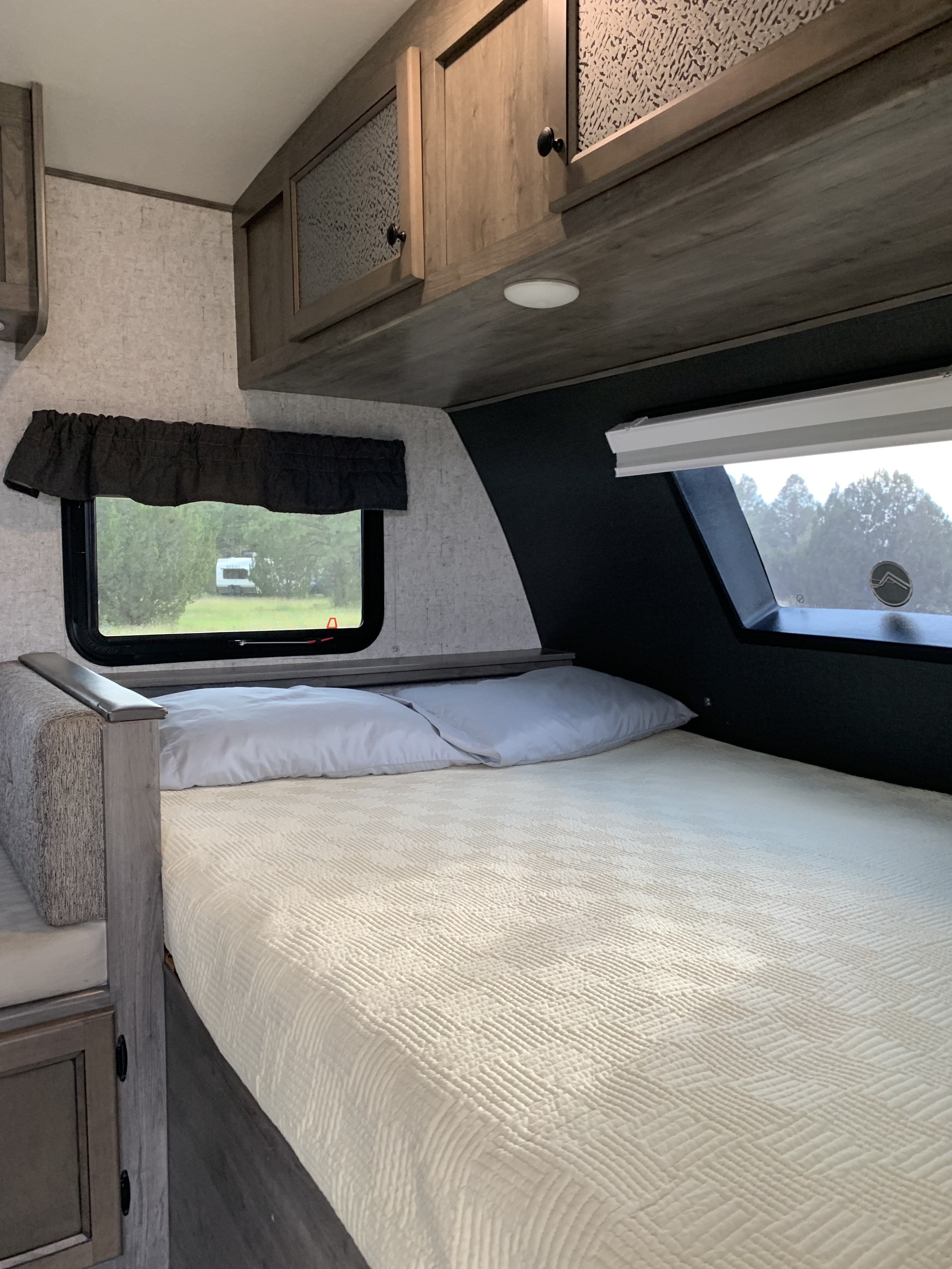 Cozy RV rental bedroom featuring a comfortable bed, overhead cabinets, and windows with scenic outdoor views.. COACHMEN NANO 2020